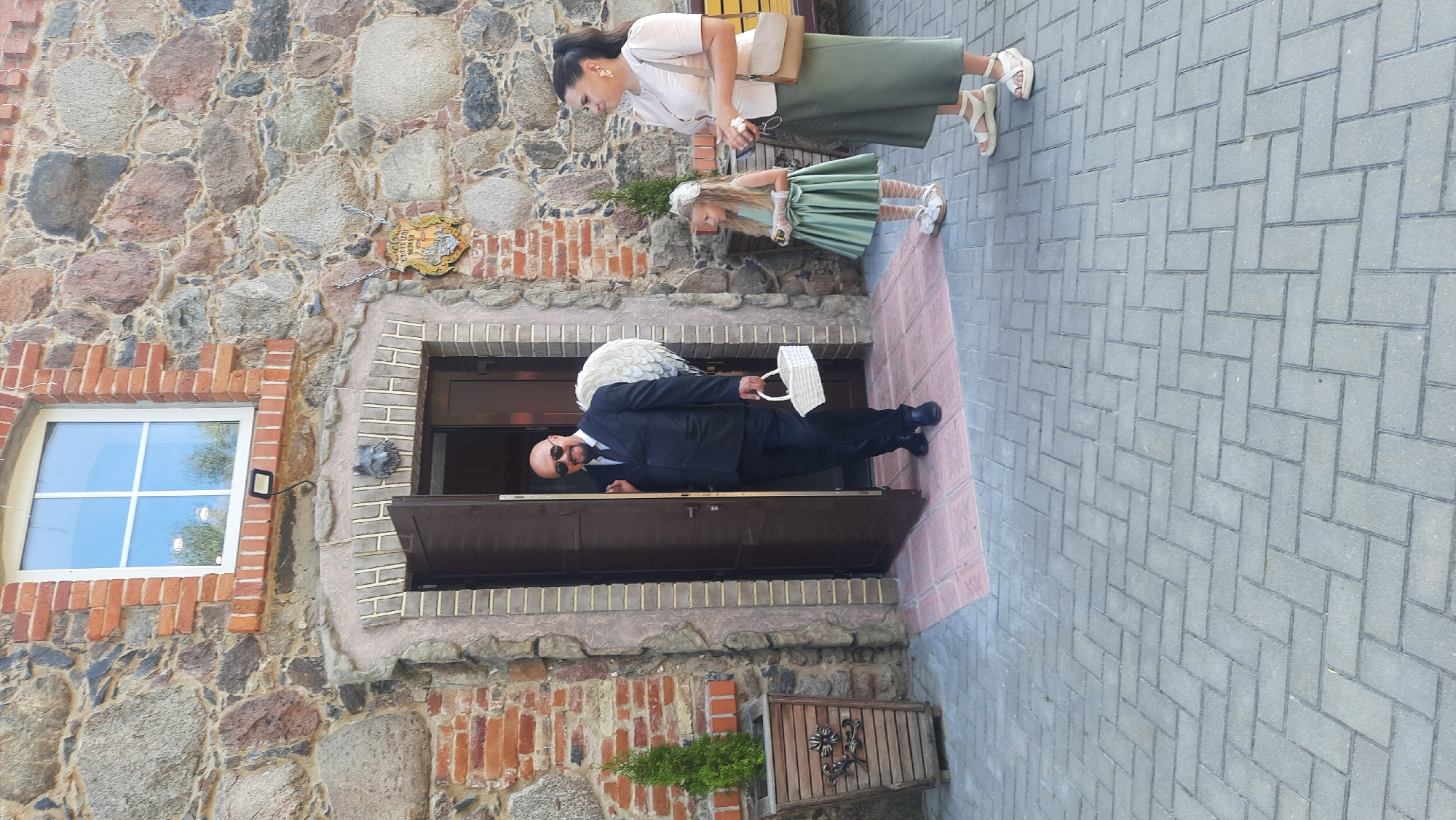 An outdoor daytime shot shows a group of people near the entrance of a stone and brick building. The building features a large window with a white frame and a stone archway above a double door. A man in a dark suit and sunglasses, with white wings attached to his back, stands in the open doorway holding a white basket. To his right, a woman in a light-colored top and olive green skirt stands with a tan purse slung over her shoulder. Beside her, a young girl in a green dress and hat looks towards the man. The ground in front of the building is paved with grey interlocking bricks. A wooden planter with a small evergreen tree is to the left of the doorway. A decorative sign is mounted on the stone wall to the right of the entrance. The scene is likely in Жодзішкаўскі сельскі Савет, Belarus, given the architectural style and potential local context.