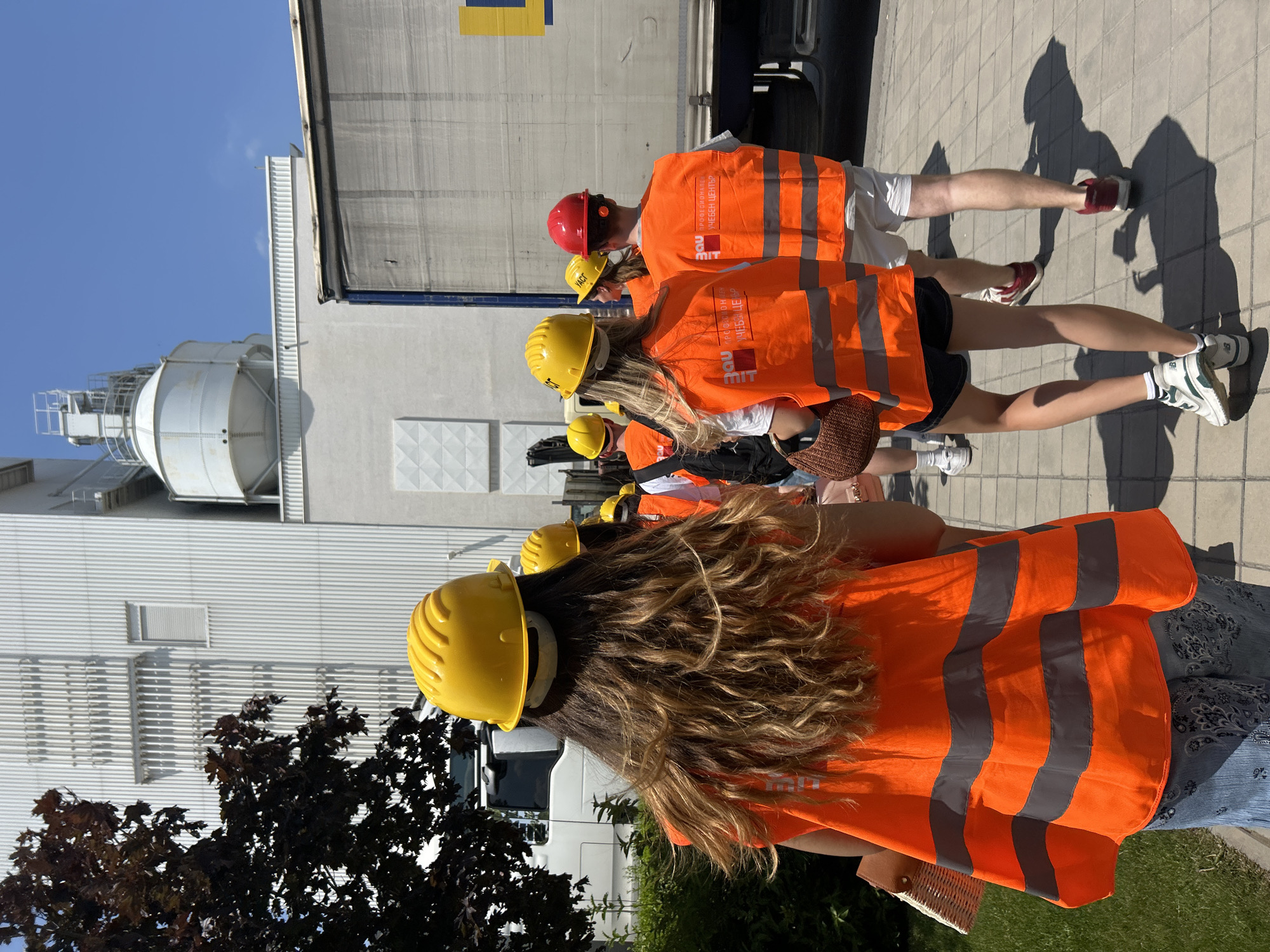 In Elin Pelin, Bulgaria, a group of people, some wearing yellow and red hard hats and bright orange high-visibility vests, are walking along a paved path next to a large industrial building. The building has a white, corrugated exterior and appears to be a manufacturing or processing facility, with a silo visible on top. A large white truck with a gray tarp-covered trailer is parked to the right of the path. The sky is clear and blue. The people are mostly facing away from the camera, suggesting they are either touring the facility or on a work assignment. The scene conveys a sense of industrial activity and safety awareness due to the protective gear worn by some individuals.