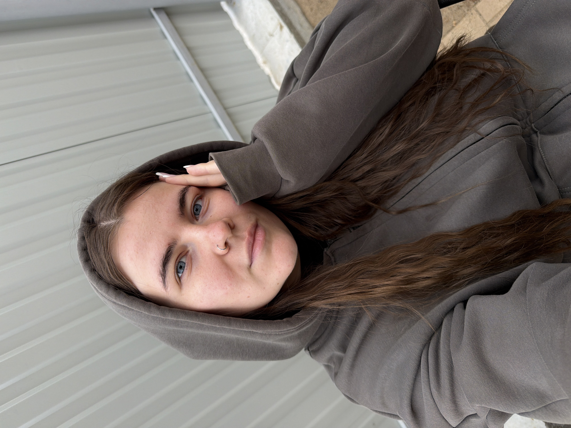 A young adult female is depicted in an outdoor setting, located in Chaikovsky, Russia. She is wearing a dark grey or olive green hooded zip-up sweatshirt with a ribbed cuff visible on her left sleeve. Her long, dark brown hair cascades over her shoulders. She has blue eyes, full eyebrows, and a silver hoop piercing in her left nostril. Her facial expression is neutral. Her left hand, with white-tipped nails, rests gently against her left temple. The background features a light grey corrugated metal surface with diagonal lines, likely a wall or roof, and a rough-textured, light-colored concrete structure is partially visible in the lower right portion of the frame. The scene is illuminated by soft, diffused light.