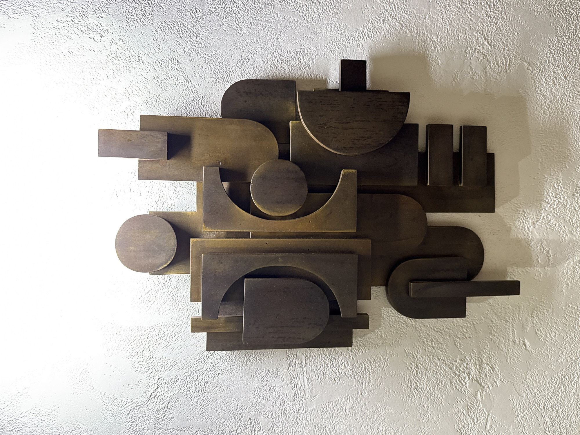 An abstract, three-dimensional wall sculpture, crafted from dark, metallic geometric shapes, is prominently displayed against a textured, off-white wall. The artwork is a complex arrangement of overlapping and interlocking elements, including circles, semicircles, rectangles, and various curved forms, creating a layered and dynamic composition. The material, appearing to be aged bronze or a similar dark metal, features a subtle, brushed finish.
Illumination from the left highlights the sculpture's contours and casts distinct, dark shadows onto the wall, emphasizing its depth and three-dimensionality. The wall itself possesses a rough, stucco-like texture. The scene is static, devoid of any human presence or activity. No text is visible on the sculpture or the wall. This indoor setting, with its textured wall, could be found in locations such as Kuta Selatan, Indonesia.