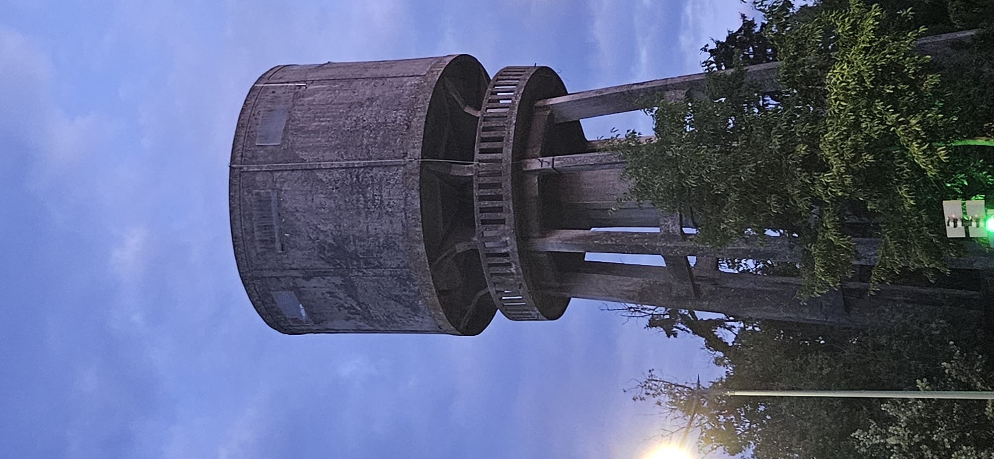 The image shows a tall, cylindrical concrete water tower located in Hévíz, Hungary. The tower has a wide, circular base supported by four thick, angled concrete pillars, creating open spaces between them. A circular walkway with a railing and vertical posts encircles the tower approximately one-third of the way up from the base. The upper portion of the tower is a large, cylindrical tank with several rectangular windows set into its concrete surface. The tower appears weathered and shows signs of age. Lush green trees surround the base of the tower, with some branches reaching up towards it. In the lower left corner, a lamppost with a bright light is visible, and in the lower right corner, a traffic light with a green signal is present. The sky above is a deep blue with scattered clouds, suggesting it is either dawn or dusk.