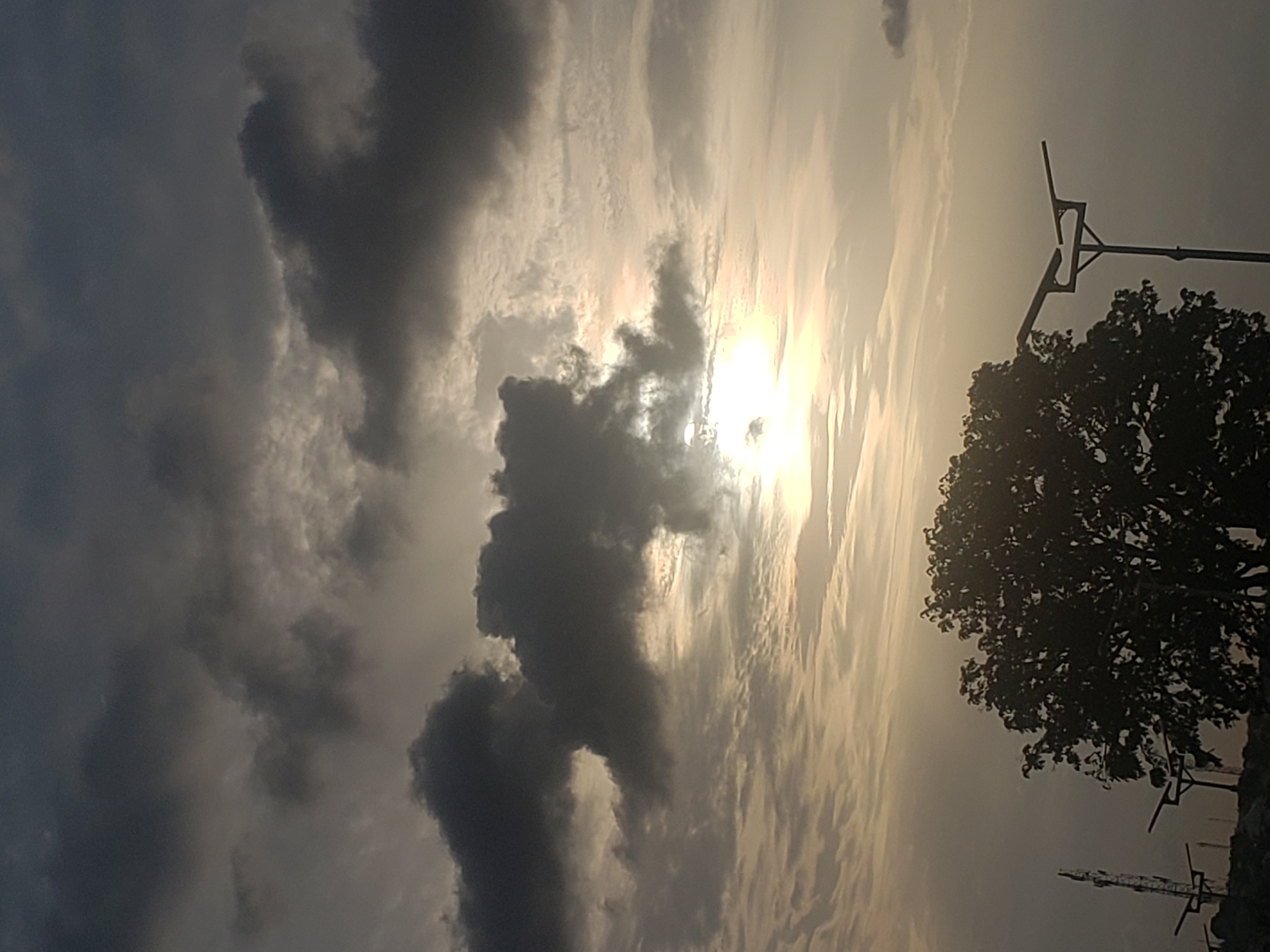 The image shows a daytime sky with dark clouds obscuring the sun. The location appears to be Adankolo, Nigeria, suggested by possible power lines and foliage silhouettes that are common in the area. The sky is a mix of dark, stormy clouds and lighter, sunlit areas. A tree is partially visible on the right side of the frame. There are no people present.