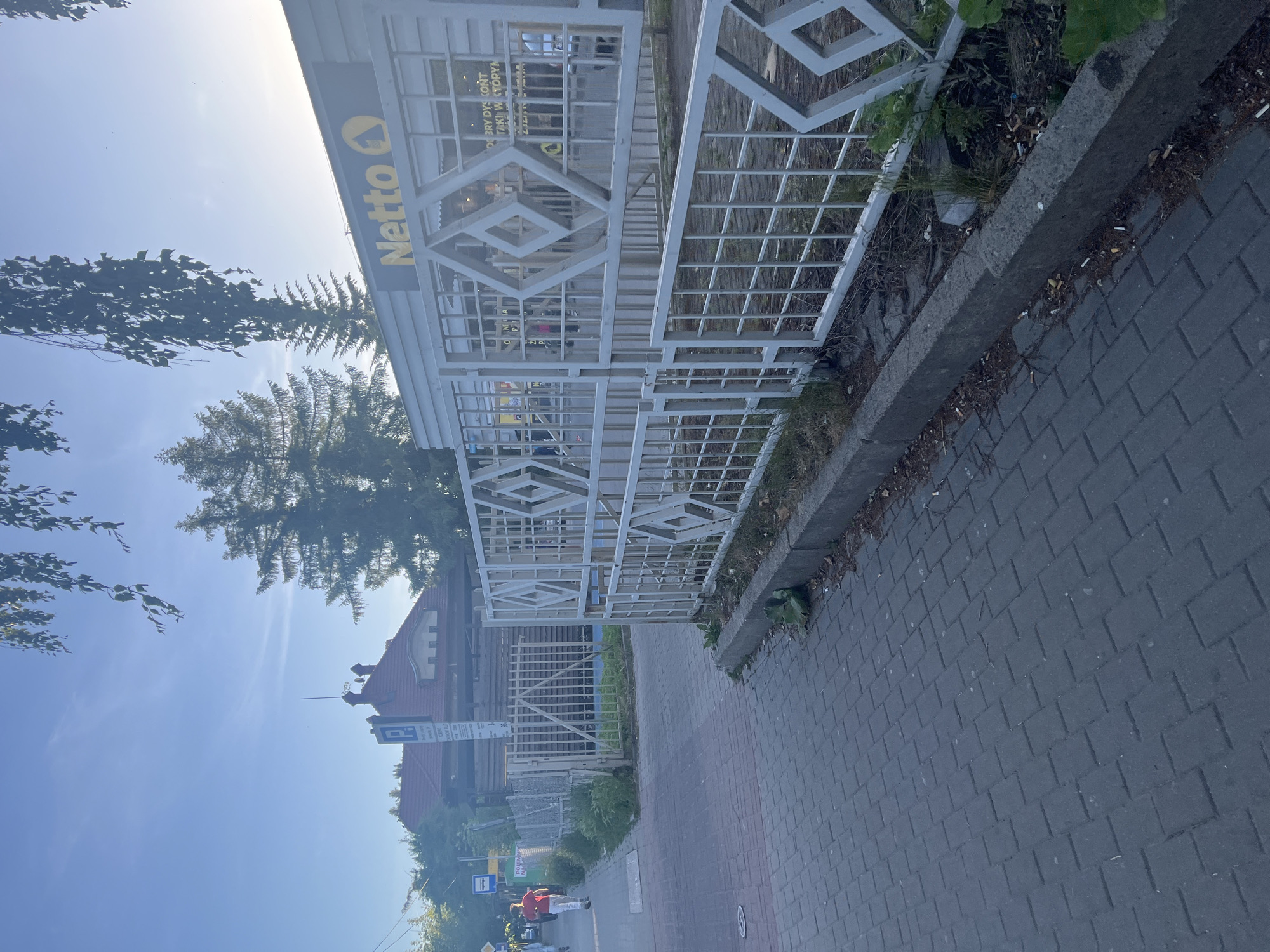 The image depicts a Netto store in an unknown city. A white fence runs along the sidewalk in front of the store, and people are walking on the sidewalk. The sky is blue, and there are trees in the background. The ground is paved with grey bricks.