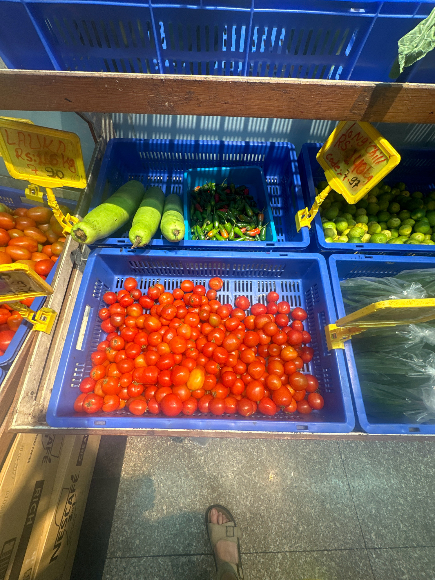 The image depicts a street-level market scene in Lalitpur, Nepal. In the foreground, a person's feet wearing sandals are visible, standing on a grey tiled surface. The main focus is on multiple blue plastic crates filled with fresh produce.
On the upper shelf, a yellow sign with "LAUKA Rs: 165 Kg 90" is attached to a crate containing several large, light green bottle gourds. To the right of the gourds, a crate holds a pile of small, vibrant green chilies mixed with some red ones. Another yellow sign above this crate reads "LONG CHILLI Rs: 140 Kg Code: 7.97". Further to the right, a crate is filled with small, light green limes or similar citrus fruits.
On the lower shelf, to the left, a crate is overflowing with small, red cherry tomatoes. Next to this, a larger crate is partially visible, also containing red tomatoes. To the far right on the lower shelf, a blue crate is filled with bundles of green onions or leeks wrapped in clear plastic.
Behind the crates, wooden shelving is visible, and a hint of sunlight casts striped shadows, suggesting an outdoor setting. In the bottom left corner, cardboard boxes with visible text like "NESCAFÉ" and "RICH" indicate a commercial area. The overall impression is a bustling local marketplace.
