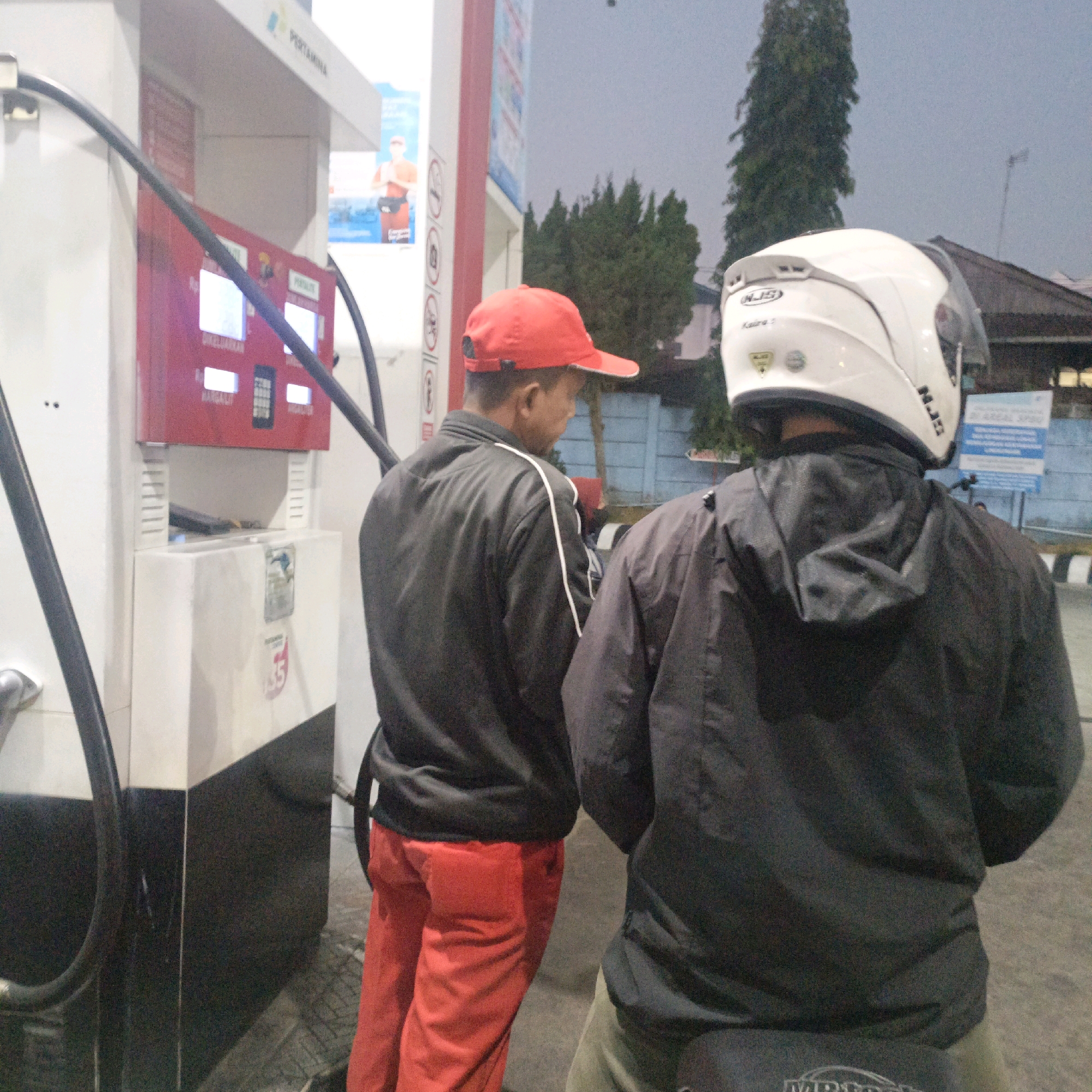 Two individuals are standing at a Pertamina gas station in Salatiga, Indonesia. The individual on the left, facing away from the camera, is wearing a red baseball cap and a black jacket with white trim, and red pants. They are standing next to a red gas pump. The individual on the right, also facing away, is wearing a white motorcycle helmet and a dark jacket, and is partially obscured by the first individual. The scene appears to be outdoors, possibly at dusk or night, with the gas station lights illuminating the area. There are trees and buildings visible in the background.