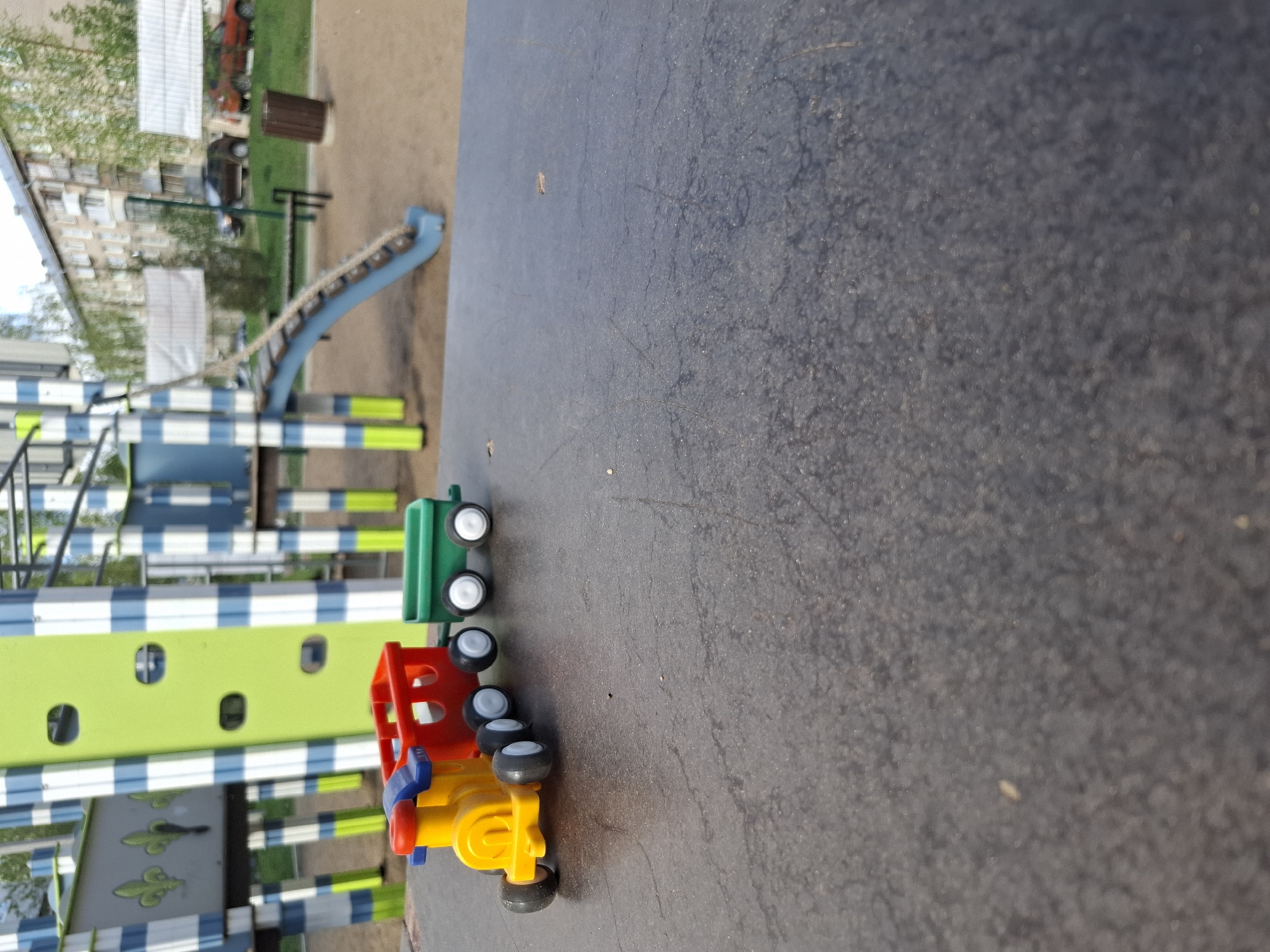 A low-angle, rotated perspective photograph captures a colorful toy truck set on what appears to be a playground surface in Chaikovsky, Russia. The toy is comprised of three detachable parts in green, red, and yellow plastic, each equipped with black wheels. The playground is characterized by blue and white striped structures and a green panel featuring circular cutouts. In the background, a slightly blurred slide structure leads to a sandy area, with a residential building partially visible. The time of day appears to be daylight, with natural lighting illuminating the scene. No people are visible in the image.