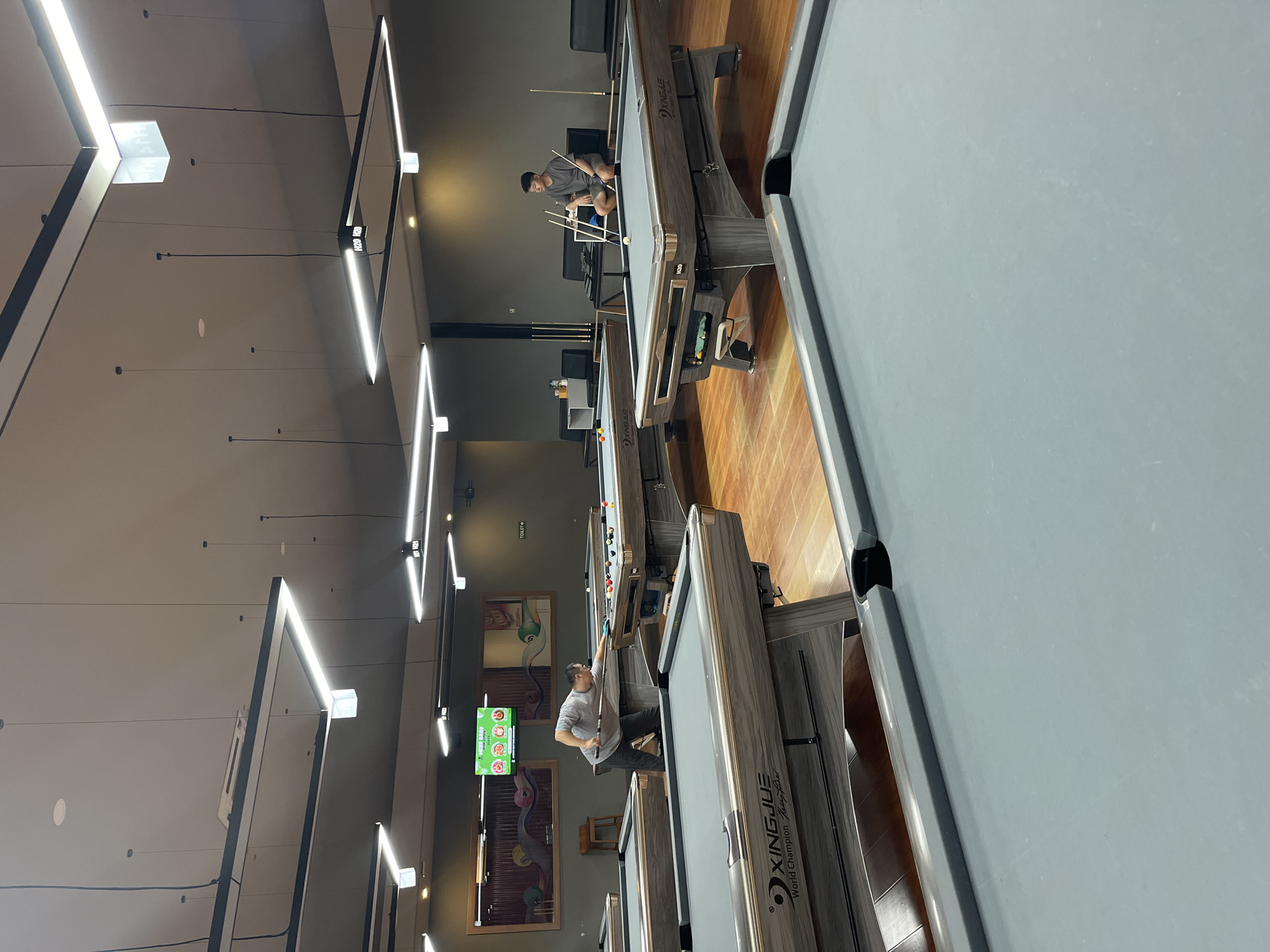 This media file shows an indoor view of a pool hall. The scene is dominated by multiple pool tables with grey felt surfaces and dark wooden frames. The tables are arranged in a way that suggests a commercial establishment.
There are at least two people visible, actively engaged in playing pool. In the foreground, a person wearing a grey t-shirt and dark pants is leaning over a pool table, cue in hand, preparing to strike a ball. Another person is visible further back, also playing pool. The lighting in the room appears to be artificial, with prominent rectangular light fixtures hanging from the ceiling, some of which are emitting bright white light. The walls are a dark, neutral color, and the floor is made of polished wooden planks.
On one of the walls, there are two framed pieces of artwork depicting cartoonish characters. A green digital screen displaying graphics is also visible on the wall, possibly indicating scores or game information. A sign pointing towards a "TOILET" is also visible. The presence of multiple pool tables, the artificial lighting, and the general ambiance suggest that it is likely daytime, or the establishment is open for business regardless of the time of day. The overall mood appears to be casual and recreational. The pool tables have branding that reads "XINGJIUE World Champion".