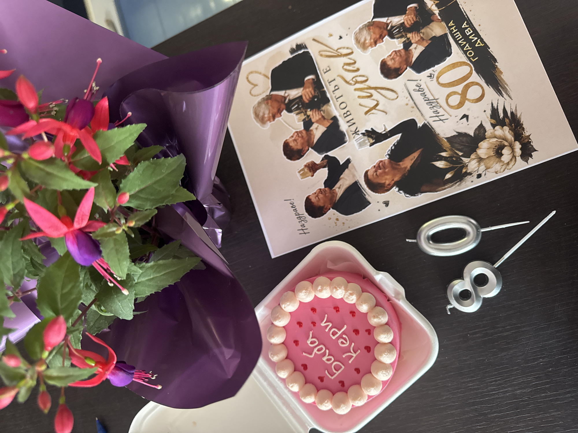 A high-angle shot captures a celebratory arrangement on a dark, textured surface reminiscent of wood grain. The composition includes a bouquet of fuchsia flowers wrapped in shiny purple material, positioned on the left side of the frame. To the right of the bouquet is a small, square cake in a white container, decorated with a pink frosting base and small white balls around the edge, adorned with tiny red hearts. The cake has text written in a script that appears to be in a Slavic language. Adjacent to the cake is a greeting card with an image of a man in various poses, also with text in a Slavic language and the number "80" prominently displayed. Numeric candles shaped as "8" and "0" are laid out to the right of the cake. The setting is likely indoors. There is no indication in the image that directly identifies the location as the city of Burgas, Bulgaria.
