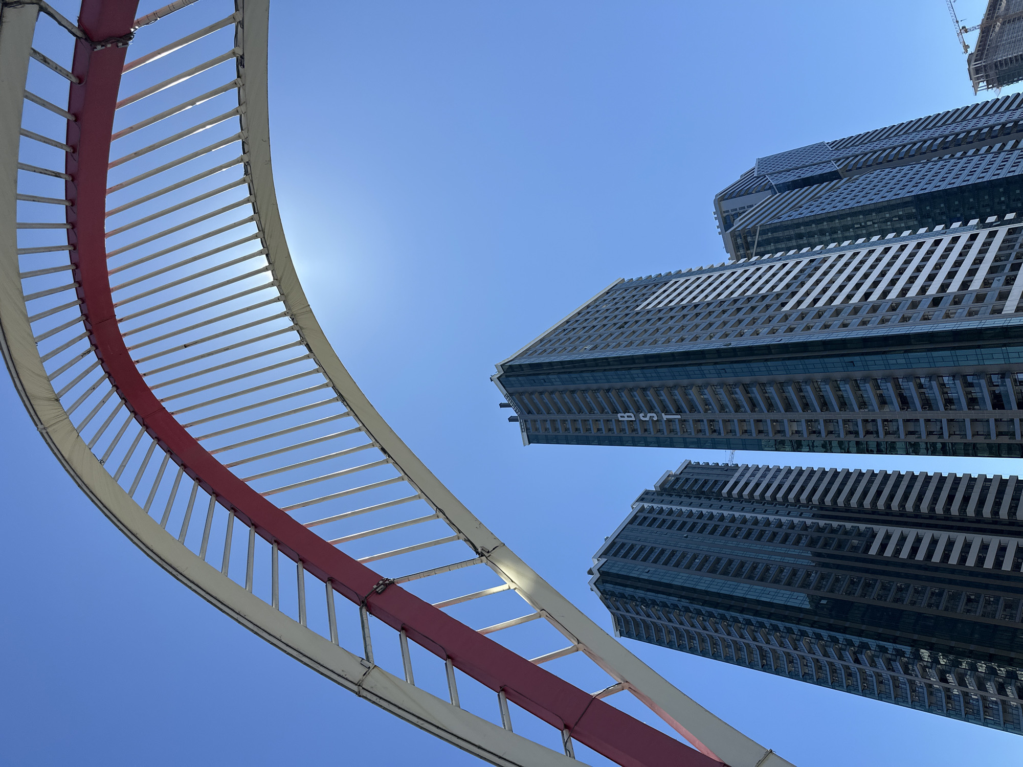 The image shows a low-angle view of three tall, modern buildings against a clear blue sky, with a partial view of a large, curved, architectural structure in the foreground.
The buildings are high-rise structures with repeating patterns of windows and balconies.
The architectural structure is a large, curved frame with a red central beam and white, spaced horizontal bars. The sun is behind the structure, creating a slight glare.
Based on the architectural style of the buildings and the overall urban landscape, this scene is very likely located in the city of Batumi, Georgia.