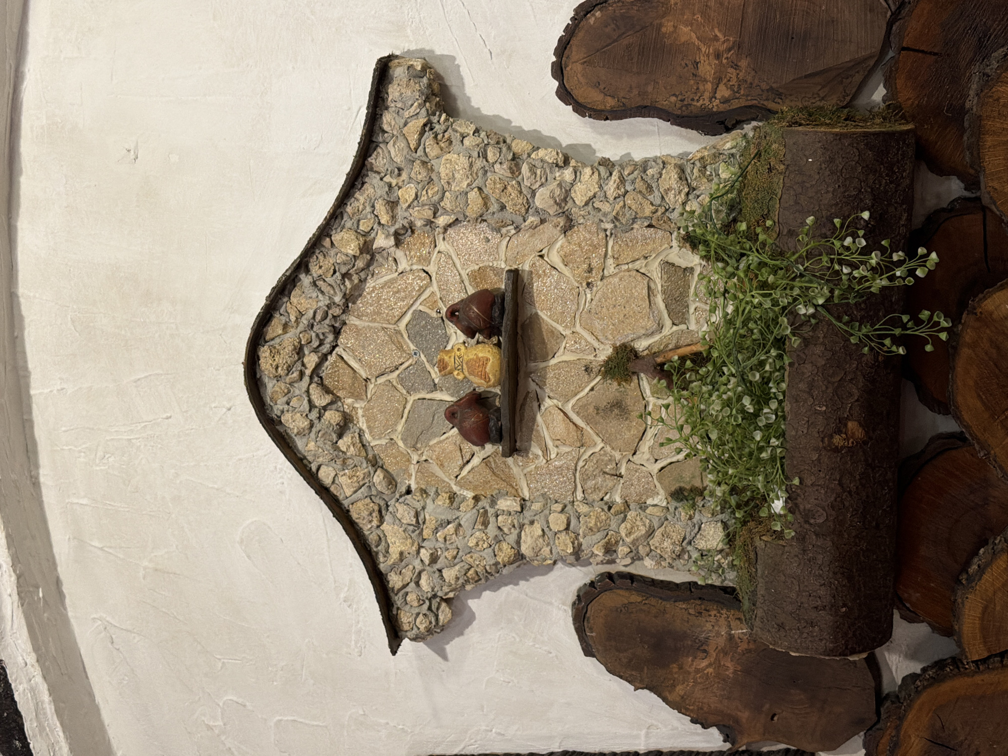 The image depicts a wall decoration, likely indoors. The decoration is an asymmetrical shape, resembling a simplified house or a rustic signboard, predominantly composed of light beige stones and mortar. A darker border outlines the shape. Central to the decoration are two small, dark brown objects, possibly ceramic or wooden figurines, positioned vertically and flanking a horizontal wooden element.
To the right of the stone-covered area, a section of dark bark-covered logs is present, with a small bush featuring tiny white flowers attached. Two segments of split wood are placed on either side of the logs.
The background consists of a textured white wall. The scene is strongly indicative of handcrafted or artisan work, possibly found in a decorative setting.
The provided location in Varna, Bulgaria, suggests this is likely a detail of an interior design feature in a business or home showcasing local craftsmanship. Without further details, it is impossible to discern its specific location, purpose or any specific interaction occurring.