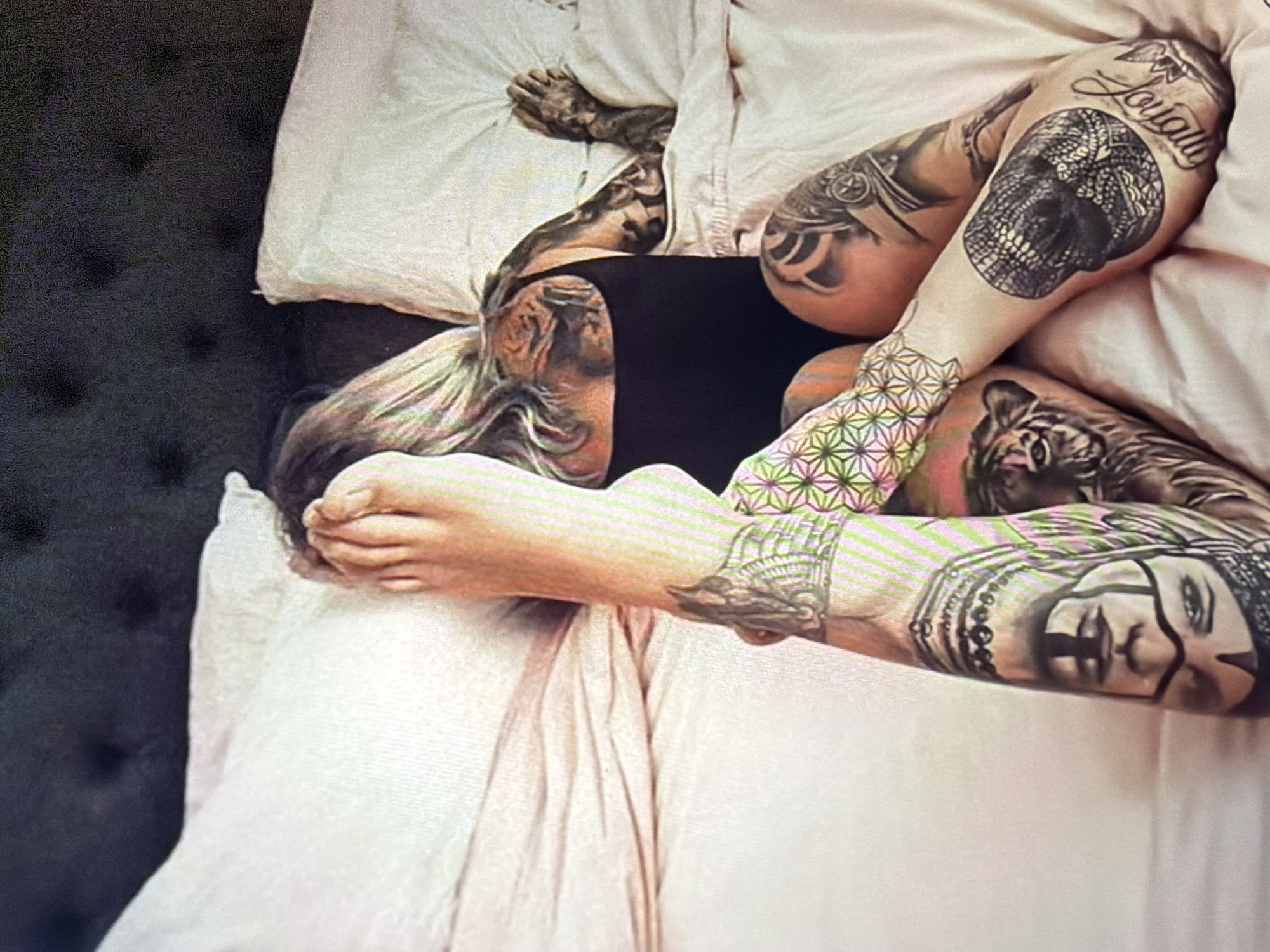 A person with extensive tattoos is lying on a bed, with their legs extended and crossed. The person is wearing a black garment. The bed has white sheets and pillows. The background shows a dark, tufted headboard. The image is taken from a slightly elevated angle. There is no indication of the city of Shumen, Bulgaria in the image.
