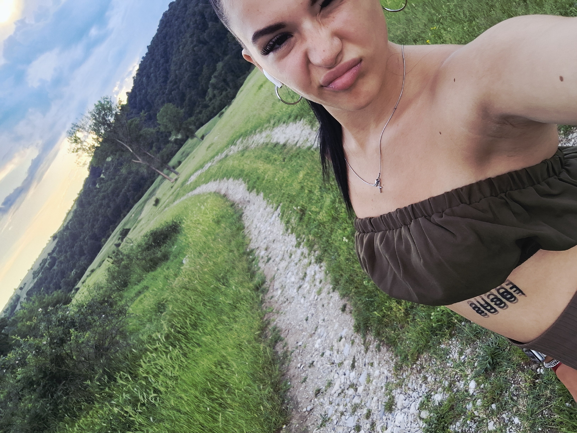 A young woman takes a selfie outdoors in a lush, hilly landscape, characteristic of Krapets, Bulgaria. She is positioned in the foreground, holding the camera at an upward angle, resulting in a tilted perspective of the scene. The woman wears a dark brown strapless top and a matching lower garment, with a silver necklace featuring a small cross pendant. She has silver hoop earrings and a white wireless earbud visible in her left ear. Her dark hair is pulled back, and she makes a pouting or slightly playful, squinting expression at the camera. A prominent tattoo of several black, stylized, stacked loop or ring designs is visible on her left side, near her ribcage.
The background features a winding dirt or gravel path cutting through vibrant green grass. Beyond the path, a dense forest covers a steep hillside, suggesting mountainous or hilly terrain. The sky above is a mix of light blue and soft white clouds, with hints of warmer light near the horizon, indicating late afternoon or early evening. The overall weather appears clear and pleasant. No discernible text is visible in the image.