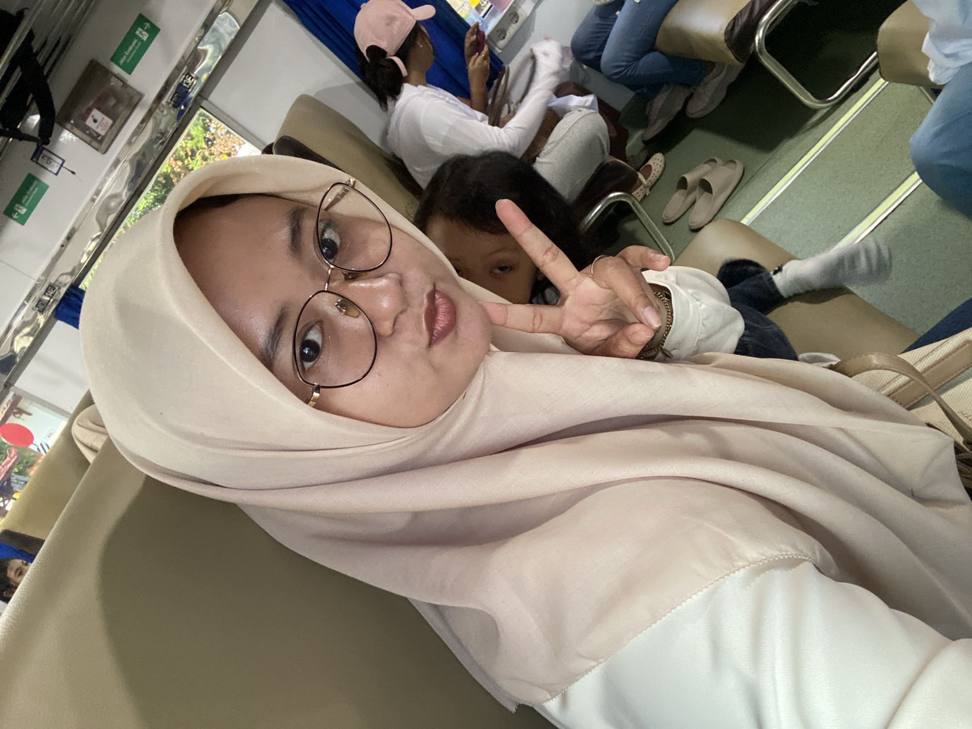 The image is a selfie taken inside a train car. The main subject is a person wearing a light-colored hijab and round glasses. They are making a peace sign with their hand. Behind them and slightly to their right is a child looking towards the camera. In the background, there are other passengers seated, including one wearing a pink baseball cap and using a cell phone. The train car has tan seats with metal frames, and the floor appears to be green. The location is likely within Indonesia, given the visual context. The city of Probolinggo is not visually confirmed based on details in the image.