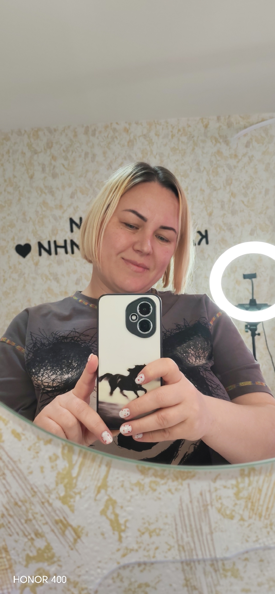 A woman takes a selfie in front of a mirror, holding a smartphone with a horse-themed case. The background includes a wall with decorative lettering and a ring light. The phone model is identified as Honor 400. No location indicators for the city of Noviy or Russia are discernable.
