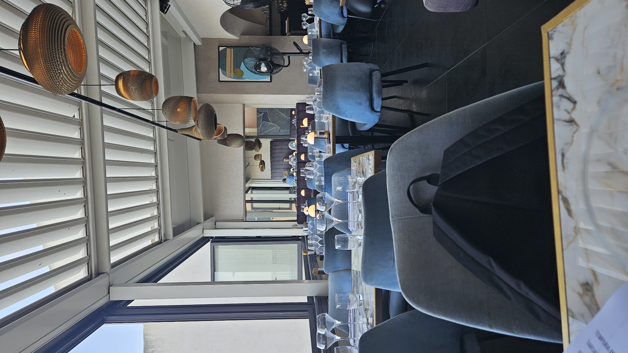 The image shows the interior of a modern restaurant with a covered outdoor seating area. The seating area is furnished with blue upholstered chairs around tables with marble-patterned tabletops. Each table is set with glassware and small lamps. In the foreground, a black backpack rests on a grey chair. The ceiling features a slatted white cover with several decorative pendant lights hanging from it. Through a large window on the left, a glimpse of an outdoor balcony and a building facade can be seen. In the background, a reflection in a mirror reveals more dining tables and a few patrons. The overall ambiance suggests a contemporary dining establishment. Based on visual cues and common restaurant styles, this scene could be located in L'Île-Rousse, France, a coastal town known for its restaurants and modern establishments.