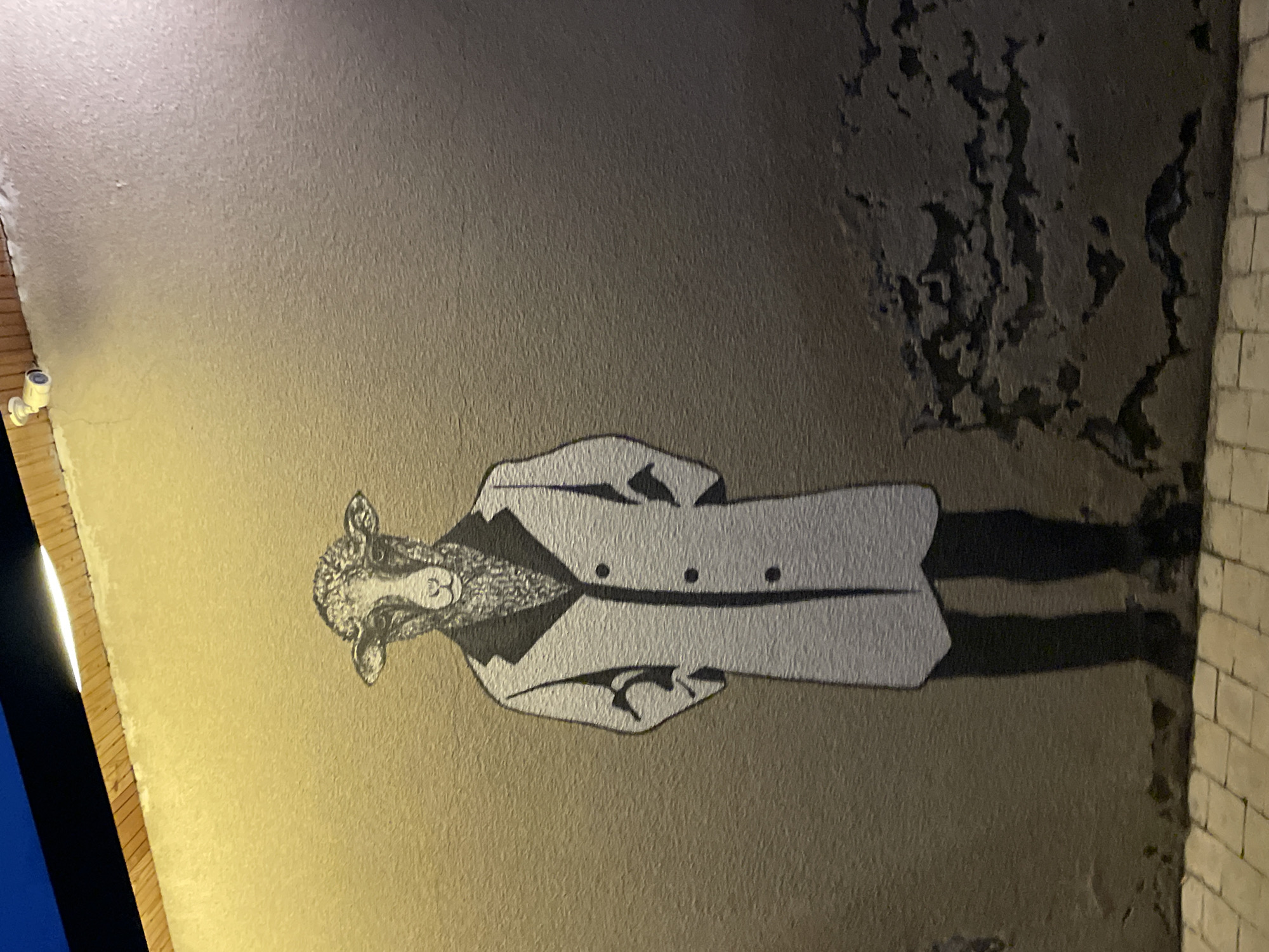 A street art mural of a sheep wearing a suit and coat is depicted on the textured wall of a building in Stepantsminda, Georgia. The mural is illuminated by a warm yellow light from above, casting shadows and highlighting the wall's rough surface. A security camera is visible on the wooden trim at the top of the wall. The lower portion of the image shows a cobblestone sidewalk in front of the wall, with the bottom edge of the mural extending to the sidewalk. The wall itself shows signs of wear and tear, with some areas of peeling plaster. The overall atmosphere is one of urban art in an outdoor setting.