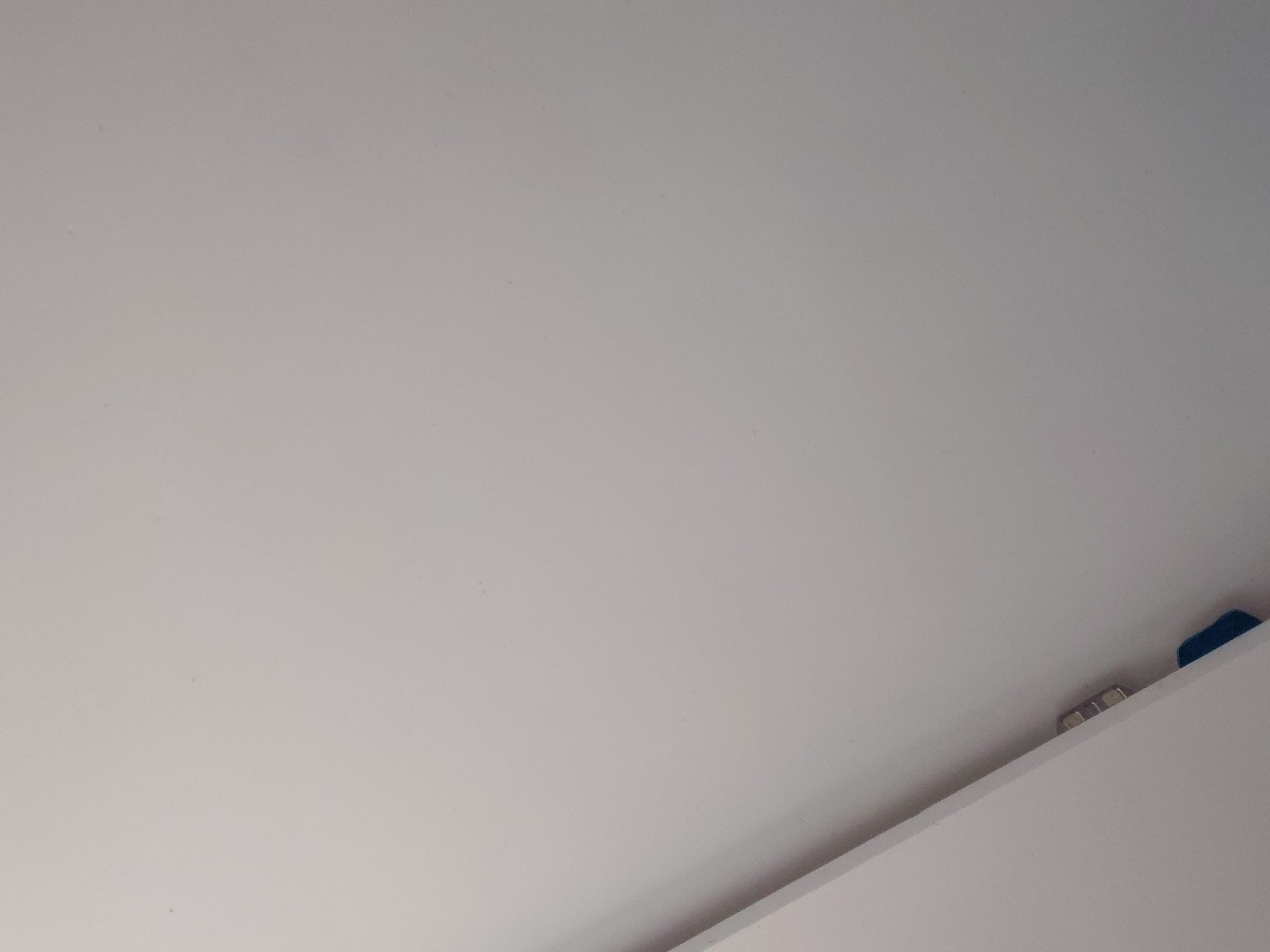 The image presents an angled, close-up view of a predominantly plain, light-colored surface, likely an interior wall or ceiling. The dominant area of the frame is filled with this uniform, off-white or light gray surface, exhibiting a subtle, almost imperceptible texture and minor variations in lighting.
In the bottom right portion of the image, a distinct, straight line indicates the edge of another surface, possibly a shelf or another wall, creating a diagonal separation from the main background. Partially obscured behind this edge are several small objects. These include a few metallic, somewhat reflective pieces, which could be fasteners, hinges, or decorative elements, appearing light and shiny. To the far right, a small section of a solid, dark blue object is visible, peeking from behind the edge.
There are no people or discernible activities taking place. The scene appears to be static and captured indoors. The lighting is diffuse and even, providing no specific indicators of the time of day or external weather conditions. No text is visible anywhere in the image. While the location context of Santa Ana, Venezuela is provided, there are no visual cues within the image itself to confirm this specific geographical location.