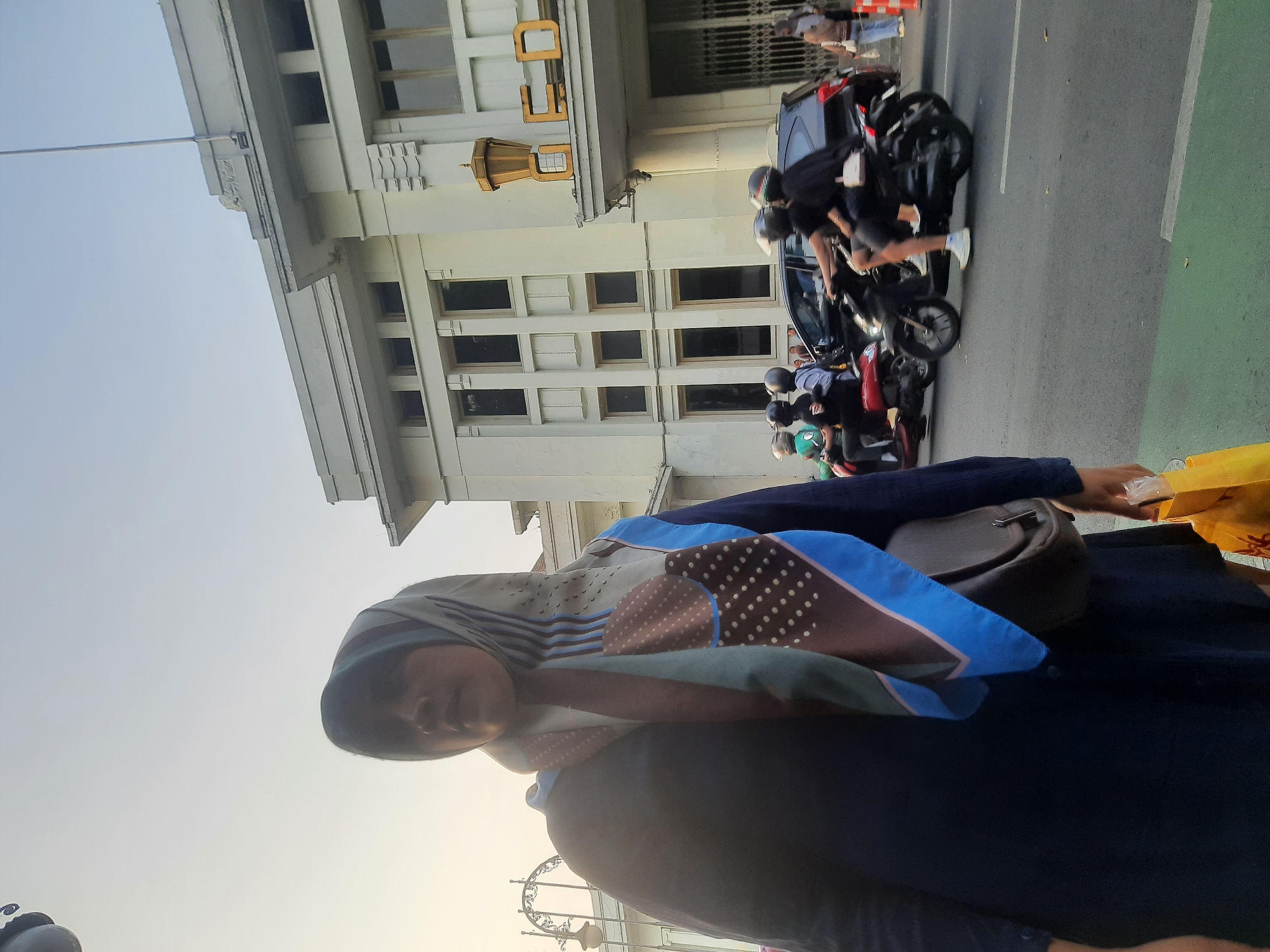 The image depicts a street scene in Bandung City, Indonesia. In the foreground, a woman wearing a dark blue dress and a patterned hijab is partially visible, holding a yellow shopping bag. She is standing on a sidewalk with a green stripe. Behind her, a light-colored building with distinctive architectural features, including rectangular windows and decorative elements, is prominent. The building appears to be an older, possibly historical, structure. A street runs in front of the building, with several motorcycles and a dark-colored car in motion. The people on the motorcycles are wearing helmets. The sky is overcast with a soft, diffused light, suggesting it might be late afternoon or early morning. The overall impression is one of urban activity and a blend of historic architecture with modern transportation.