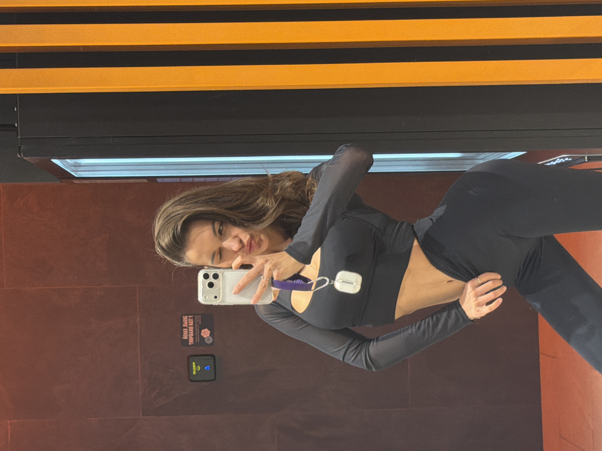 A young woman with long brown hair takes a selfie in a bathroom. She is wearing a black long-sleeved crop top and black pants. She is holding a white smartphone in her left hand, and her right hand is resting on her hip. She is winking and puckering her lips. The bathroom has reddish-brown walls and a black and orange ceiling. There is a sign on the wall behind her that reads "KONAN DRAMA SUSHI RAMEN." The bathroom is located in the city of Krasnoyarsk, Russia.