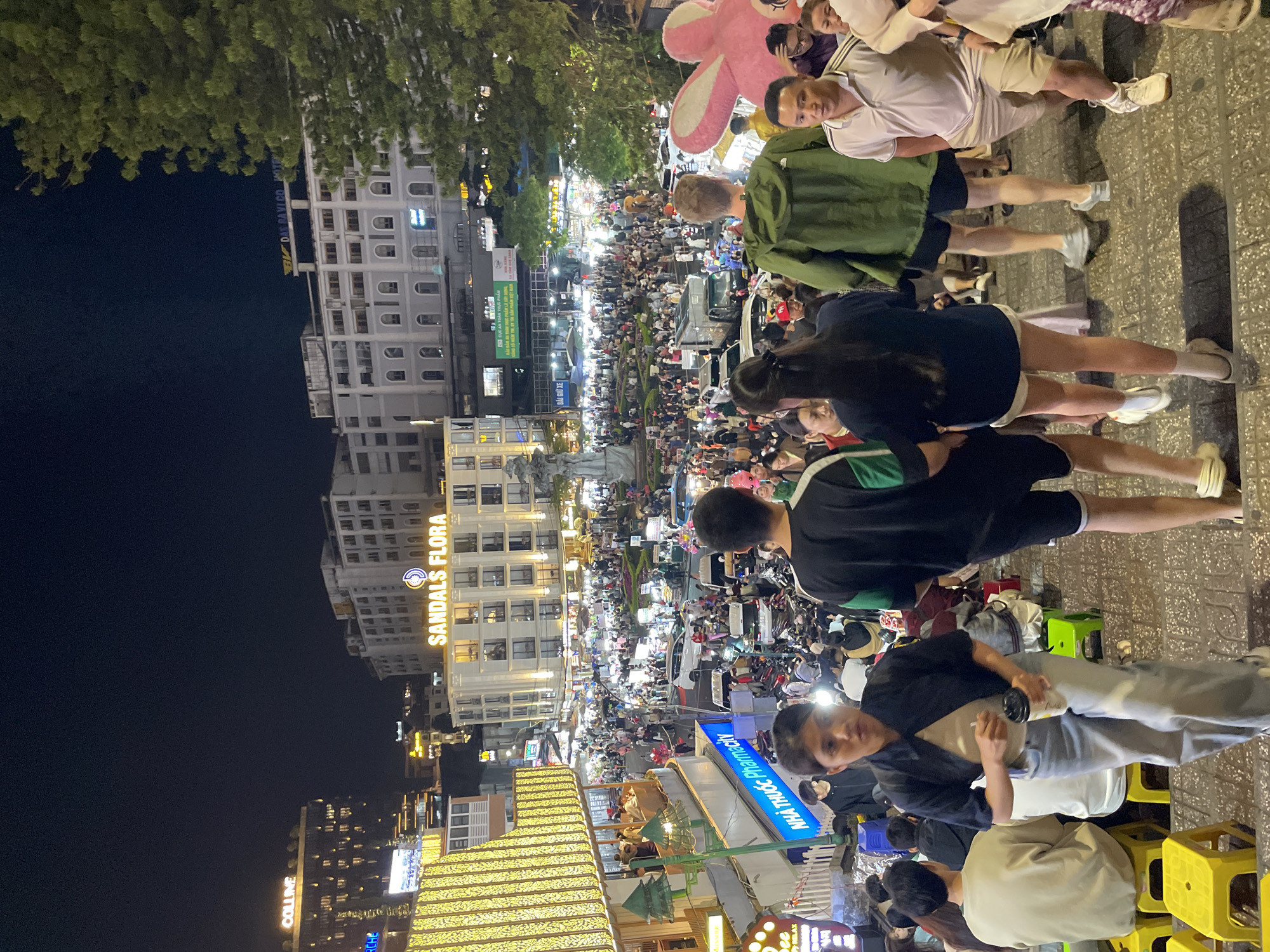 The image depicts a bustling night scene in the city of Da Lat, Vietnam, likely on Nguyen Chi Thanh street, known for its vibrant night market. A large crowd of people are seen walking along a street lined with shops and buildings, illuminated by numerous lights. The foreground shows several individuals on stone steps, with a woman in a black jacket and jeans in the center holding a drink. Behind her, more people are visible, some walking down the steps, others in the crowd below. The background reveals a dense urban environment with multi-story buildings adorned with illuminated signs, including "SANDALS FLORA". Vehicles, predominantly motorcycles, are interspersed with the pedestrians in the street. The overall atmosphere suggests a lively and crowded commercial area at night.