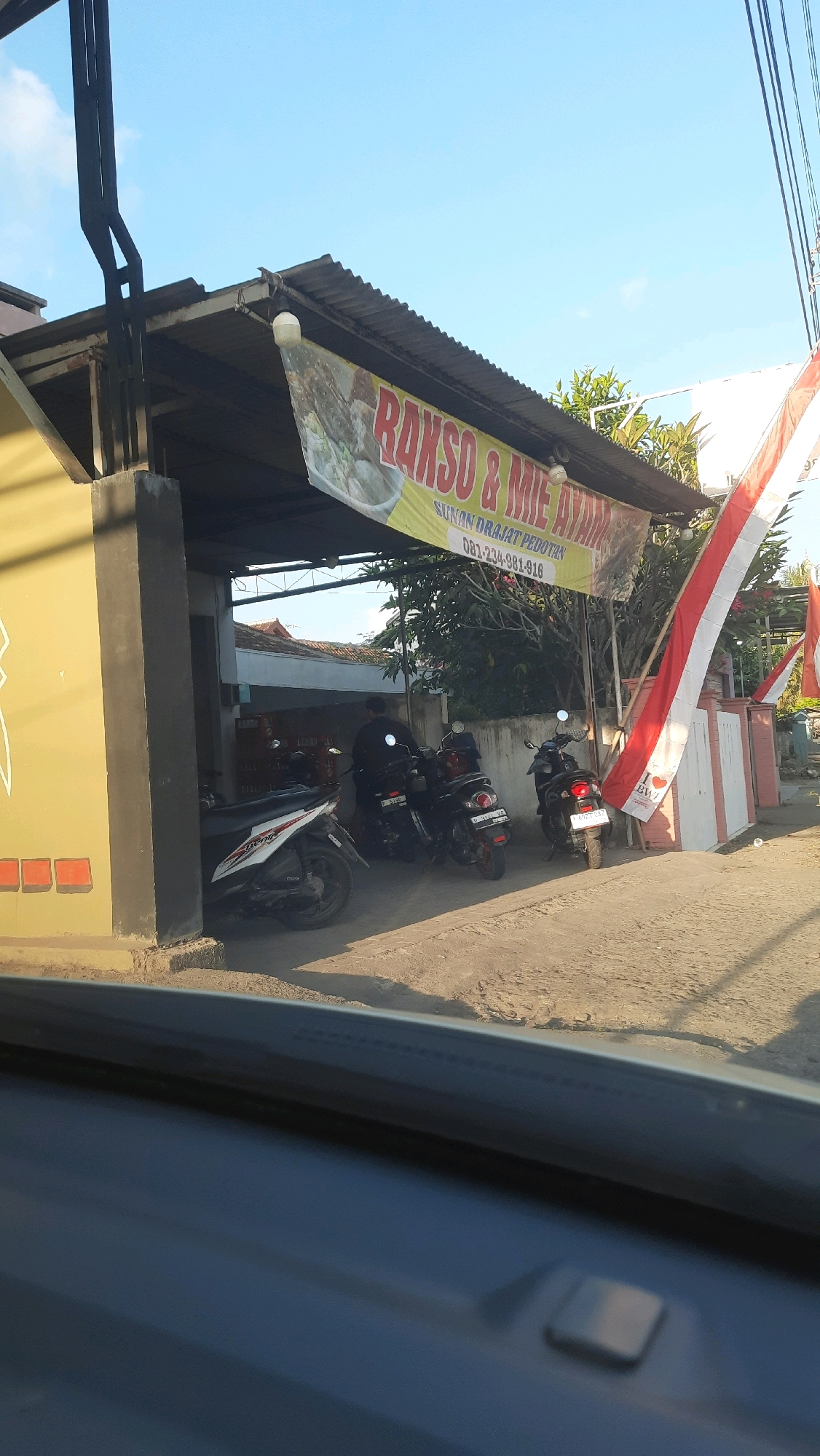 The image captures a storefront in Bangorejo, Indonesia, specifically a food establishment identified by a banner as "RAKSO & MIE AYAM." The banner also displays contact information: "081-234-981-916." The building features a yellow exterior wall and a corrugated metal roof supported by a black metal structure. In front of the establishment, several motorcycles are parked. One person is visible, standing near a black motorcycle, seemingly interacting with the establishment. Indonesian flags, red and white, are prominently displayed on poles, suggesting a national celebration or patriotic decoration. The ground is dirt and gravel, typical of an outdoor environment in the area. The sky is clear blue with a few wispy clouds. The perspective suggests the photo was taken from a vehicle, possibly looking out through a windshield.