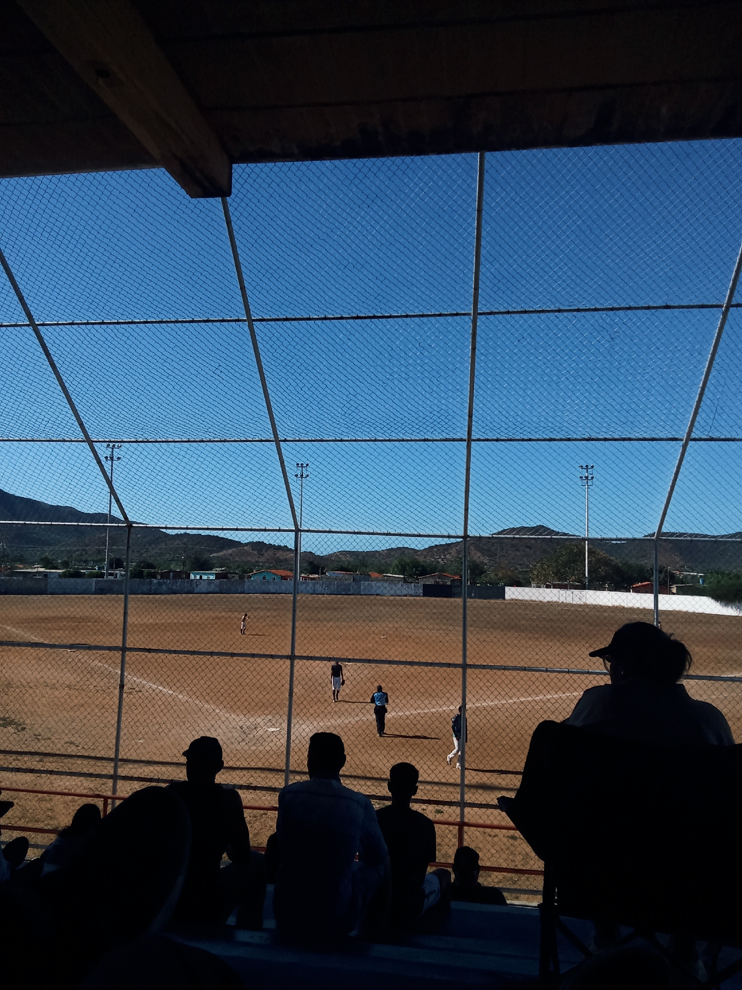 From the perspective of a spectator under a roofed structure, a baseball game unfolds on a dirt field in Juan Griego, Venezuela. A chain-link fence separates the viewer from the field. The sky is blue and clear. Light poles surround the field. In the distance, there are small hills and buildings. Silhouetted spectators sit in the foreground, watching the game. Several people are on the field.