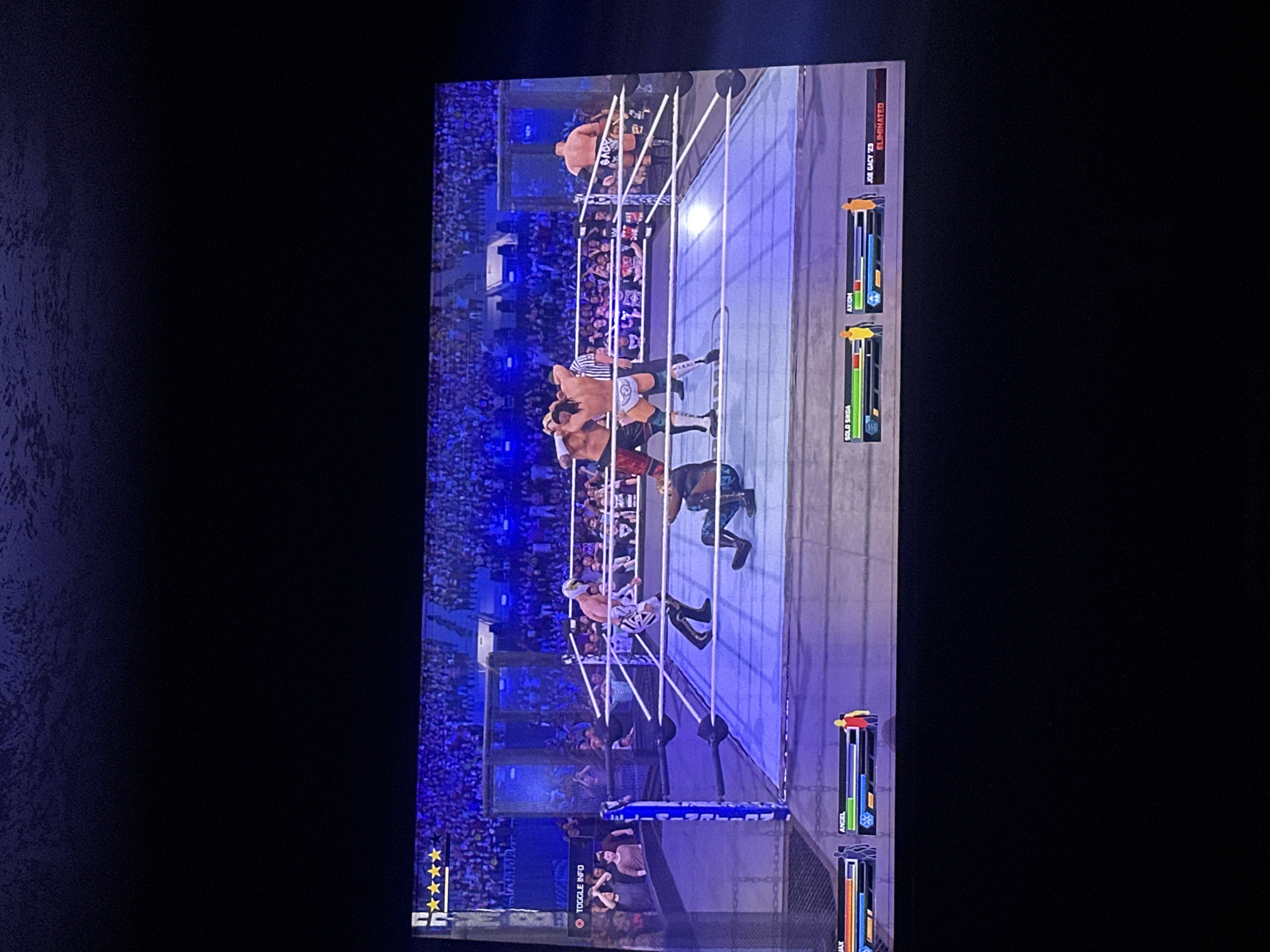 The image shows a WWE video game being played on a screen. The match is taking place inside a Hell in a Cell structure. Several wrestlers are in the ring and around the cage. The names Solo Sikoa, Axiom, and Joe Gacy '23 are visible. Joe Gacy '23 has been eliminated. The rating of the match is five stars. The location is not identifiable from the image.