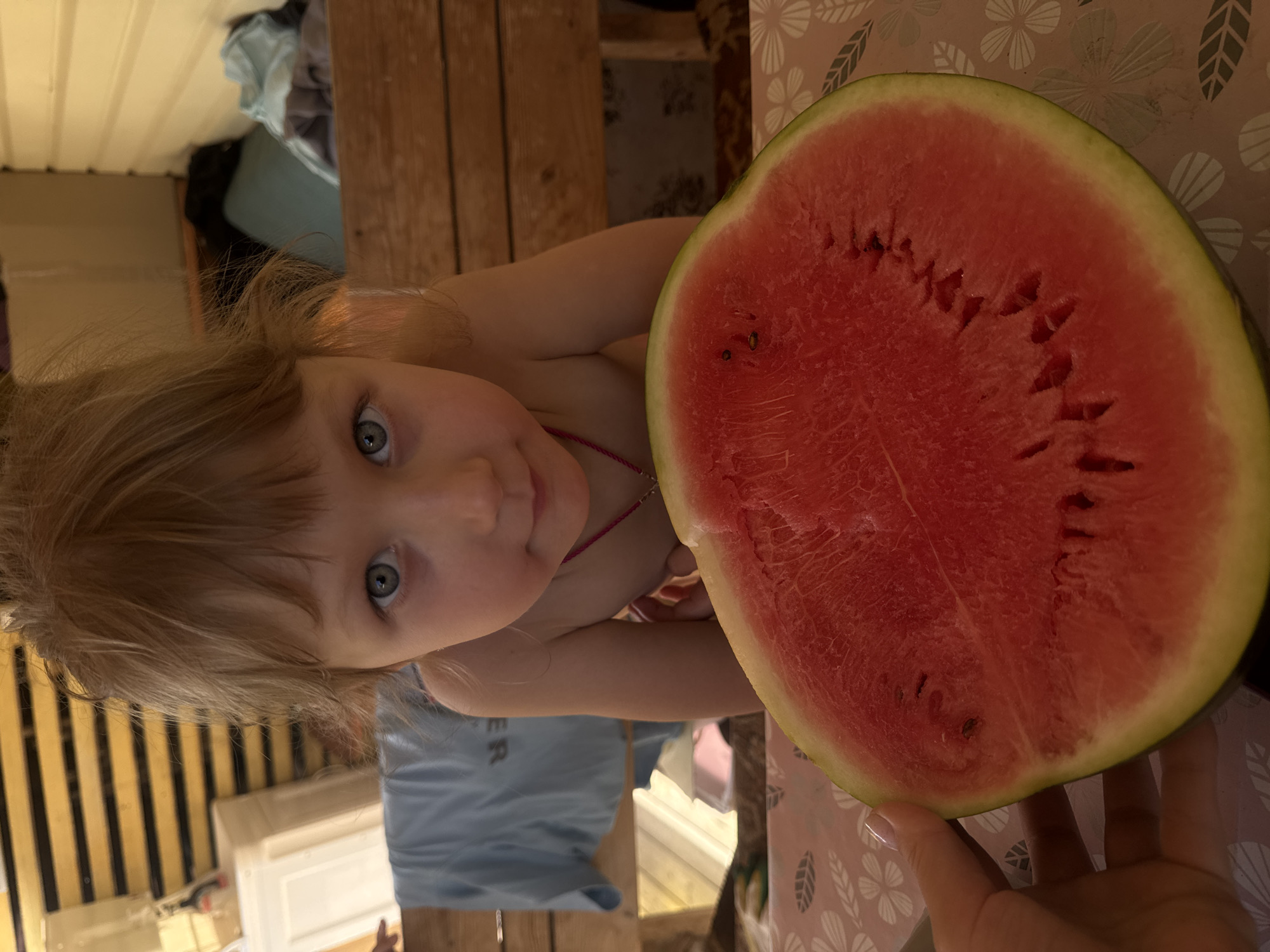 The image displays a young child and a large half-slice of watermelon, captured in a setting stated to be the city of Chaikovsky, Russia. A fair-skinned child with light hair and light-colored eyes looks directly at the camera, positioned behind the watermelon. The child appears to be bare-chested and wears a thin pink necklace. In the foreground, a prominent red watermelon half, containing numerous visible black seeds, displays a green rind transitioning to pale yellow. A light-skinned human hand with manicured nails is visible on the lower left, holding the watermelon. The watermelon and child rest upon a surface covered by a light-colored tablecloth featuring a repeating pattern of white or light grey leaves and flowers against a beige or pinkish background. The background consists of various elements including wooden slatted structures, possibly furniture components, and obscured items such as a light-colored container and folded garments, including a light blue shirt. The scene is illuminated, suggesting an indoor or sheltered outdoor environment.