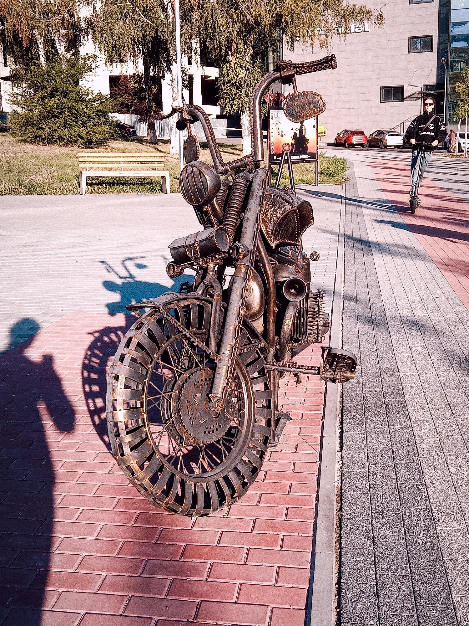 This is a medium-shot, eye-level, outdoor photograph of a metal sculpture of a motorcycle parked on a red brick pathway next to a paved sidewalk. The sculpture is detailed, featuring a large front wheel with metal slats forming the tire, a prominent engine, handlebars with mirrors, and chains. The sculpture casts a long shadow on the brick pathway. In the background, to the left, is a grassy area with a light-colored park bench and trees. To the right, a person wearing sunglasses and a black jacket is riding a kick scooter on the sidewalk. Beyond the sidewalk is a modern building with large windows and some cars parked in front. The scene is illuminated by bright sunlight, suggesting it is daytime. The location is identified as Chișinău, Moldova.