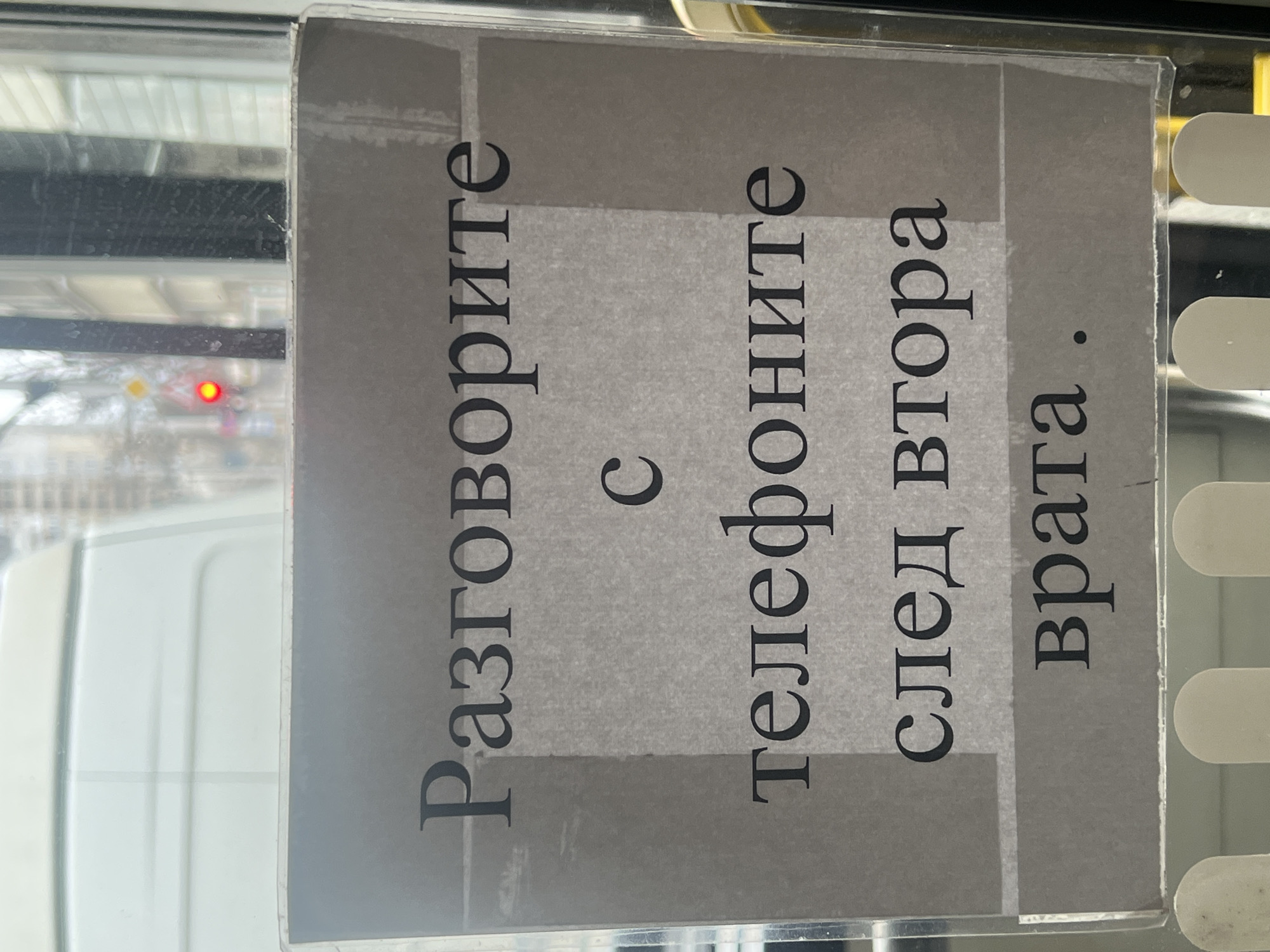 The image depicts a sign in Bulgarian script, likely affixed inside a public transportation vehicle, prohibiting phone conversations near the second door. The text reads: "Разговорите с телефоните след втора врата." This indicates the location is likely in Bulgaria, and given the presence of Cyrillic script, a major Bulgarian city like Sofia is probable. A blurry view of an urban environment is visible through the window, suggesting a city bus or tram. A white panel is partially visible, likely part of the interior of the vehicle.
