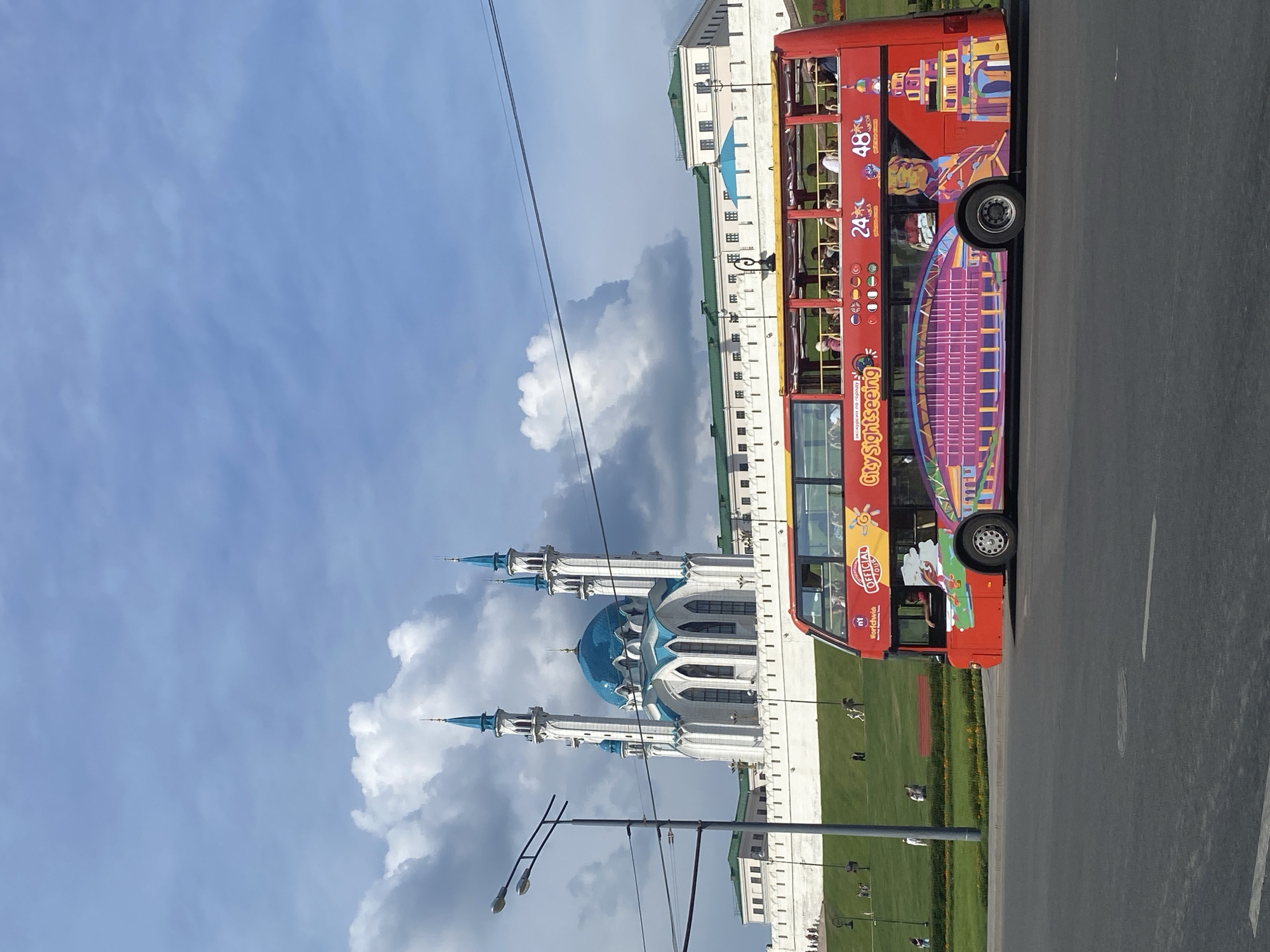 The image shows a bright red double-decker tour bus with "City Sightseeing" branding driving on a street in front of the Qol Sharif Mosque in Kazan, Russia. The mosque, a prominent landmark, features white walls, a large blue dome, and four tall, slender minarets topped with turquoise spires. Behind the mosque, a long, white walled structure with green roofs stretches across the frame. The sky above is a mix of blue and large, fluffy white clouds, with some darker grey clouds present as well. The bus is decorated with colorful images, including what appears to be a stylized depiction of a bridge and towers. Several people are visible on the upper deck of the bus, looking out at the scenery. In the background, on a grassy hill leading up to the mosque, a few individuals are walking. The street in the foreground is paved, with white lane markings visible. A lamppost is situated to the left of the frame.