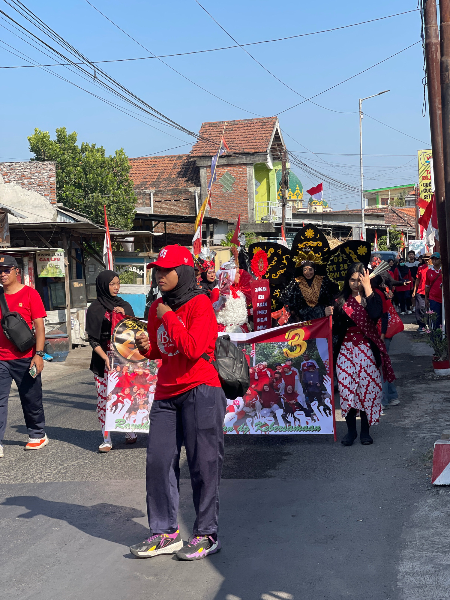 A group of people are marching in a street parade in Candi, Indonesia. The people are wearing red and white clothing, and some are carrying banners. There are buildings on either side of the street, and the sky is blue. A large banner is being carried by several people, with the number "3" prominently displayed. The banner also features an image of people in red shirts, and text that appears to be in Indonesian. One woman in the foreground is wearing a red shirt and a black hijab, and is looking towards the right. Another woman to her left is wearing a black hijab and a patterned skirt, and is holding a portion of the banner. To the left of the group, a man in a red shirt and black pants is walking. In the background, a mosque with green domes is visible. The overall scene suggests a festive or celebratory event.