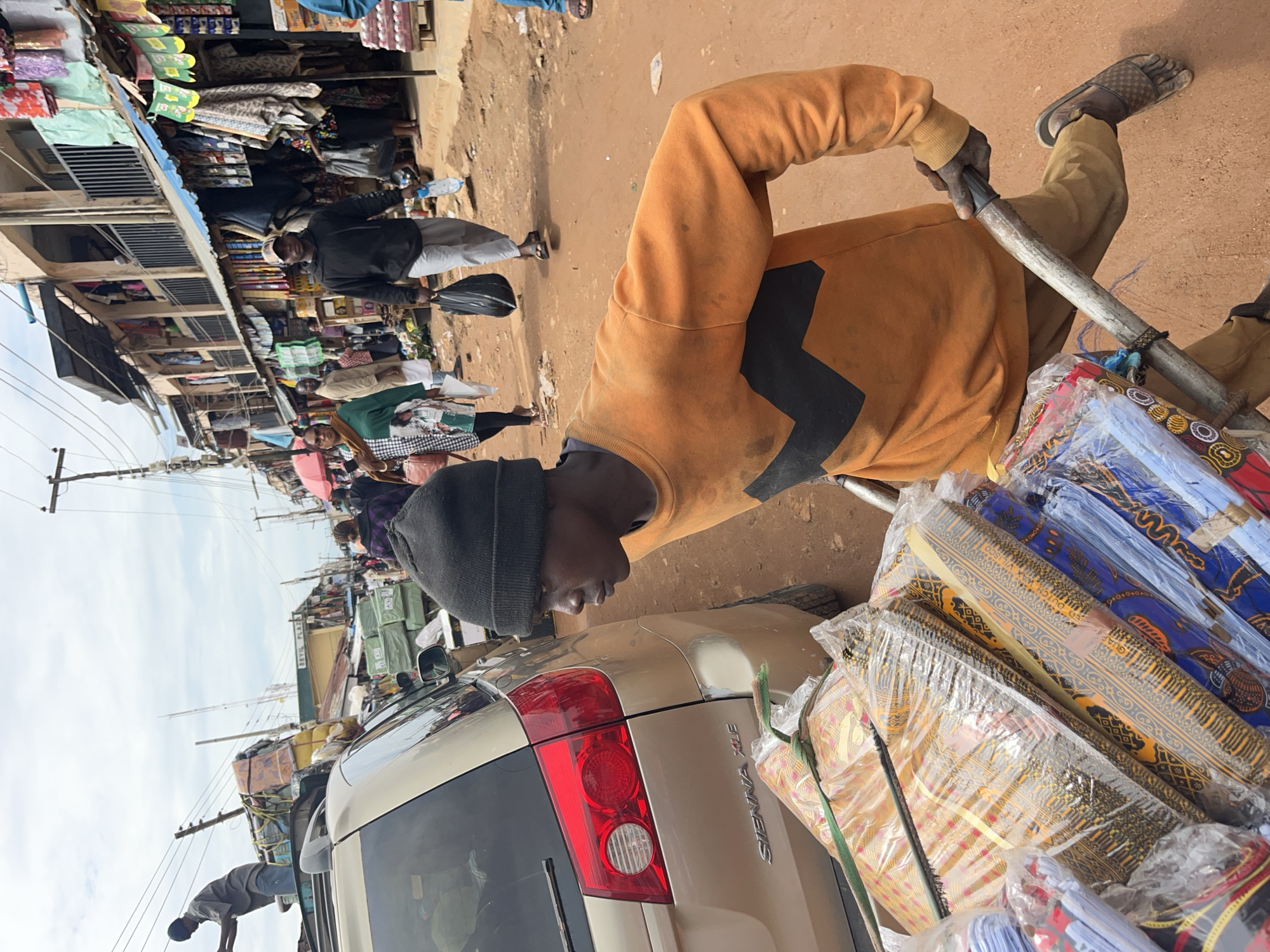 The image portrays a bustling open-air market scene in Jos, Nigeria, captured from a slightly elevated and tilted perspective. The ground consists of unpaved, reddish-brown dirt, with numerous market stalls lining both sides.
In the foreground, a young man is prominently featured, pushing a handcart piled high with goods. He is dressed in an orange long-sleeved shirt with black geometric patterns, dark pants, and a black beanie, with sandals on his feet. His left hand grips a metal pole that serves as part of the handcart's frame. The cart is loaded with numerous rolls of colorful textiles, individually wrapped in clear plastic. Many of these fabrics display vibrant, intricate patterns characteristic of African prints, with partial text like "DUTCH" visible on some rolls.
In the mid-ground and background, other individuals are visible, moving through the market or standing near stalls. One person in a black jacket and light pants is seen further back. The market stalls are semi-permanent structures, many with corrugated metal roofs and open fronts, displaying a variety of merchandise including clothing and packaged items. Green and yellow banners hang from some of these stalls.
A light-colored (likely gold or beige) minivan or SUV is partially visible in the mid-ground, with its rear section and taillight facing the viewer. The vehicle's model, "SIENNA XLE," is clearly visible on its tailgate. Overhead, multiple utility poles with a dense network of wires cut across the sky, which appears overcast or hazy, suggesting daytime hours with soft, diffuse light. The overall scene depicts a lively atmosphere of daily commerce and movement within the market.