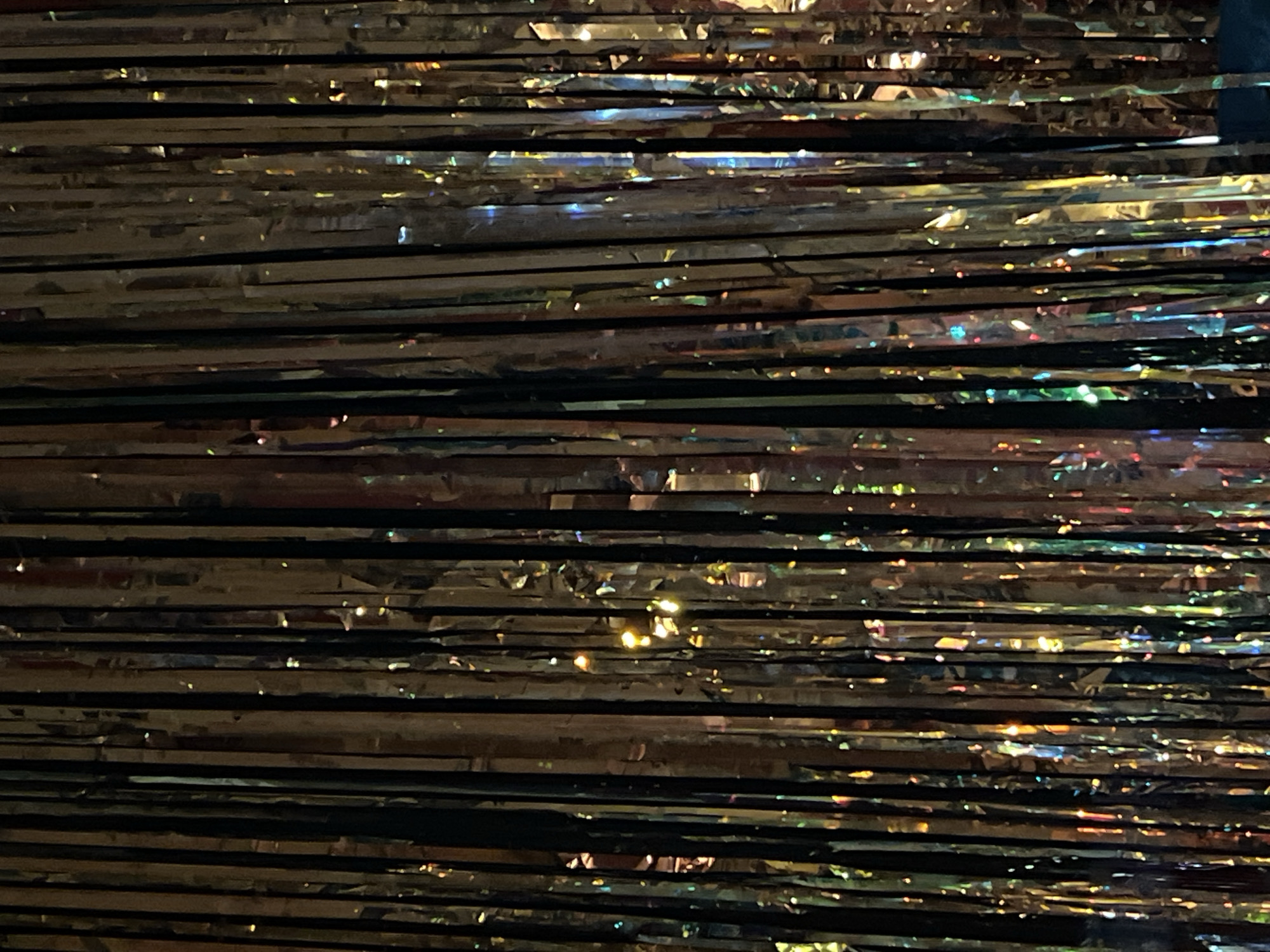 The image shows a close-up of a metallic fringe curtain. The horizontal strands of the curtain are shiny and reflective, with a dark color. The background is dark, and the lighting creates highlights and shadows on the fringe. The image shows an indoor setting. The location in the city of Palilula Urban Municipality, Serbia, cannot be determined from the image.