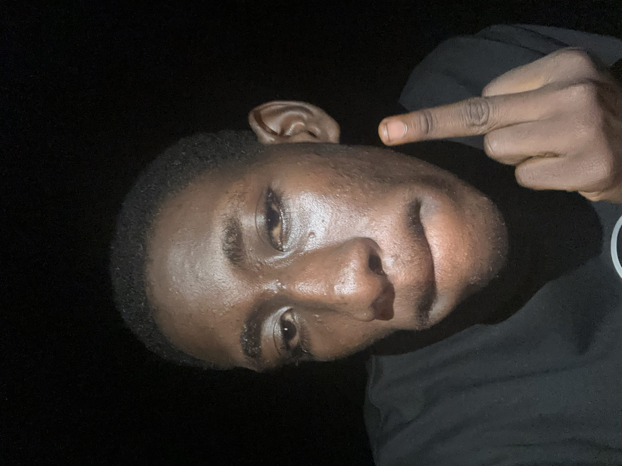 A close-up, head-and-shoulders, selfie-style portrait features a young Black individual with dark skin, looking directly at the camera. The person has short, dark hair and their face appears shiny, likely due to sweat. They are wearing a black t-shirt. The background is uniformly dark, suggesting an indoor or nighttime setting. The individual's left hand is raised, with the middle finger extended in a gesture, while the other fingers are curled. The lighting is directed towards the face, creating strong highlights and shadows. The image appears to be taken in Durimi, Nigeria, though no specific landmarks or environmental indicators confirm this location within the image itself.