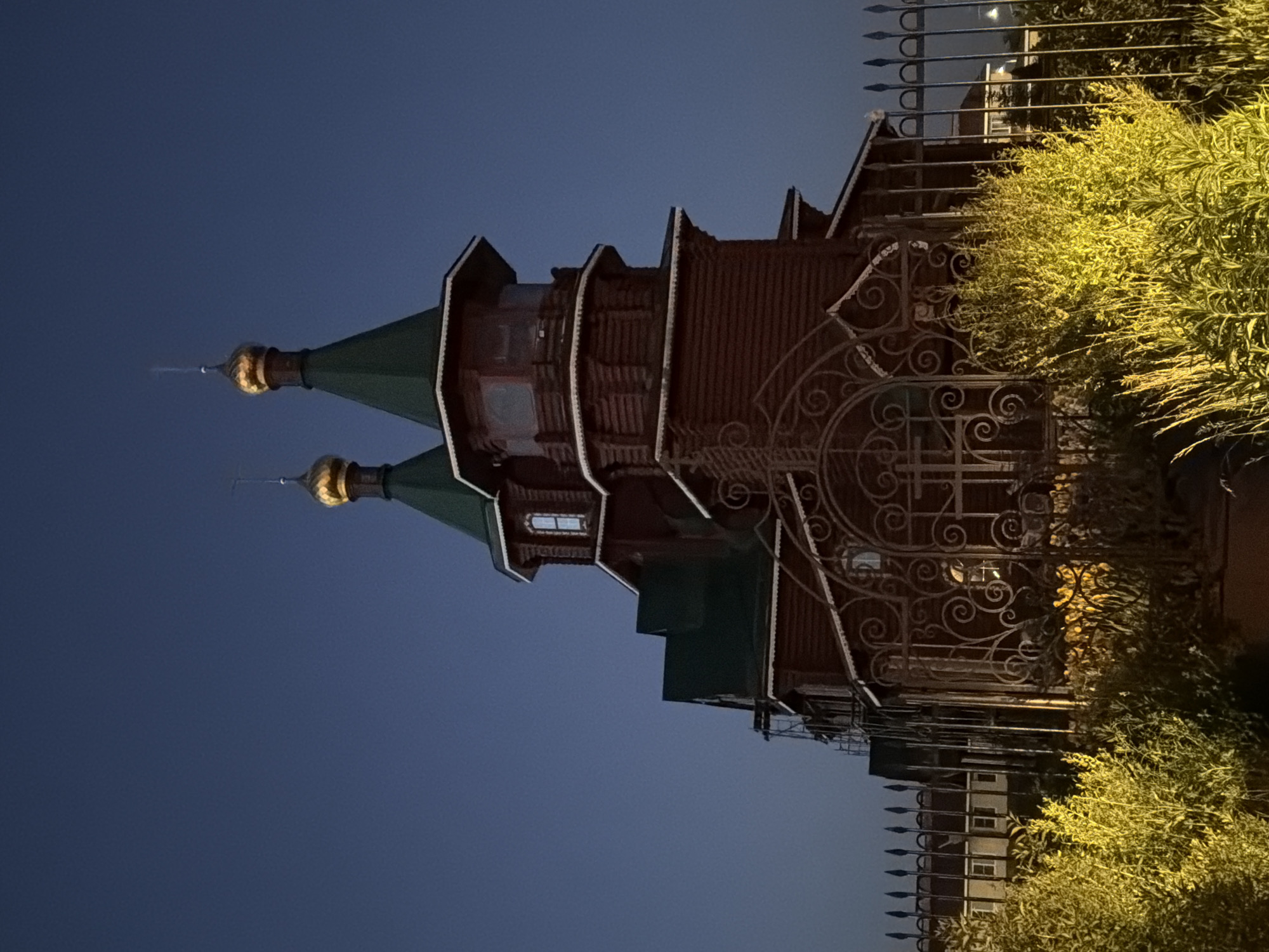 An image captured at dusk or night depicts a multi-tiered wooden building, featuring architectural elements characteristic of Orthodox churches, located in Новинки, Russia. The structure is built from dark reddish-brown logs, accented with white trim beneath the eaves and around the window frames. It is topped with dark green conical roofs and two prominent golden onion domes, each culminating in a slender spire. Small windows on the upper levels are illuminated from within, casting a soft yellow glow. A decorative wrought-iron fence with intricate scrollwork stands directly in front of the building, complemented by a simpler metal fence with pointed finials extending laterally. The lower foreground is occupied by dense, brightly illuminated yellow-green foliage. The background features a uniform dark blue sky and indistinct outlines of other buildings visible beyond the fences. Partial scaffolding is present on the left side of the main wooden structure.