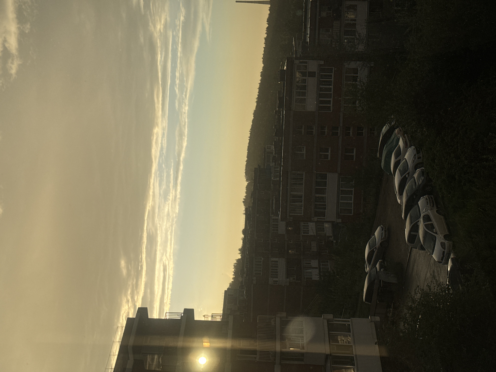 The image depicts a view of apartment buildings and parked cars against a backdrop of a hazy sunset in the city of Markova, Russia. The sun is low on the horizon, casting a warm, golden light and creating lens flare on the left side of the image, near a tall apartment building. The sky is filled with scattered clouds, some of which are illuminated by the setting sun. In the foreground, several cars are parked on a paved area between buildings, some of which are covered with tarps. Trees and dense foliage are visible in the lower portion of the image and also in the background, obscuring parts of the lower floors of the buildings. A distant smokestack or tower can be seen on the far right, silhouetted against the sky. The overall scene is one of a typical urban residential area during the golden hour.