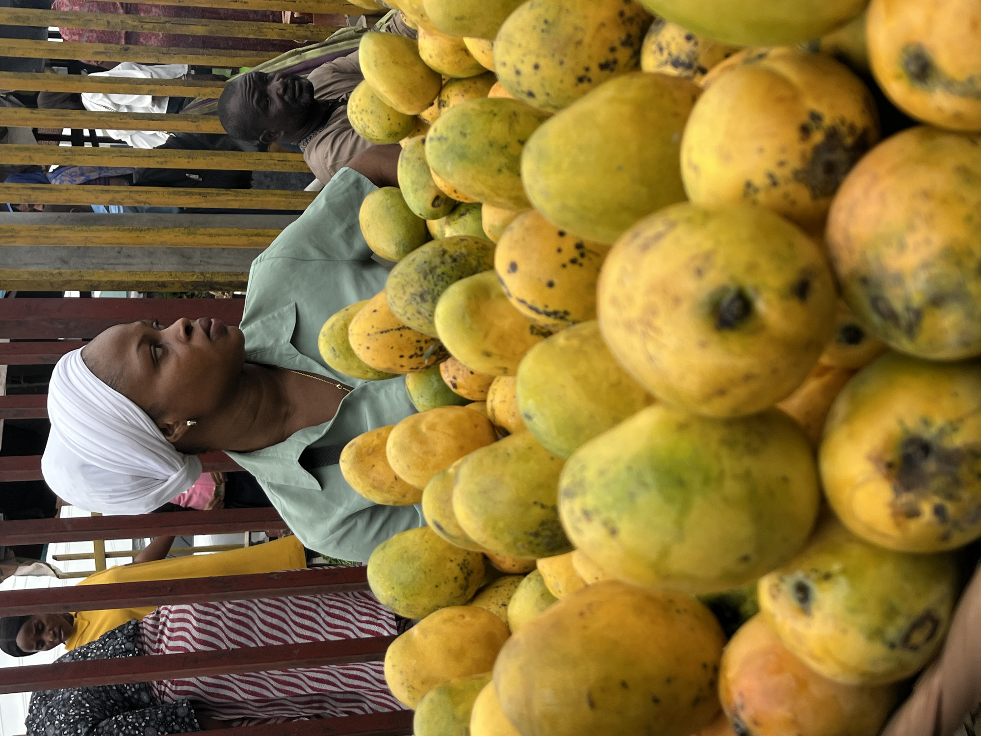 The image shows a close-up view of a large pile of yellow and green mangoes in Lekki, Nigeria. The mangoes are stacked densely. Through the metal railings behind the fruit, partial views of people are visible, including an African woman wearing a white head covering and a light green collared shirt. Another person is visible to the left, and two males are visible on the top left of the frame.