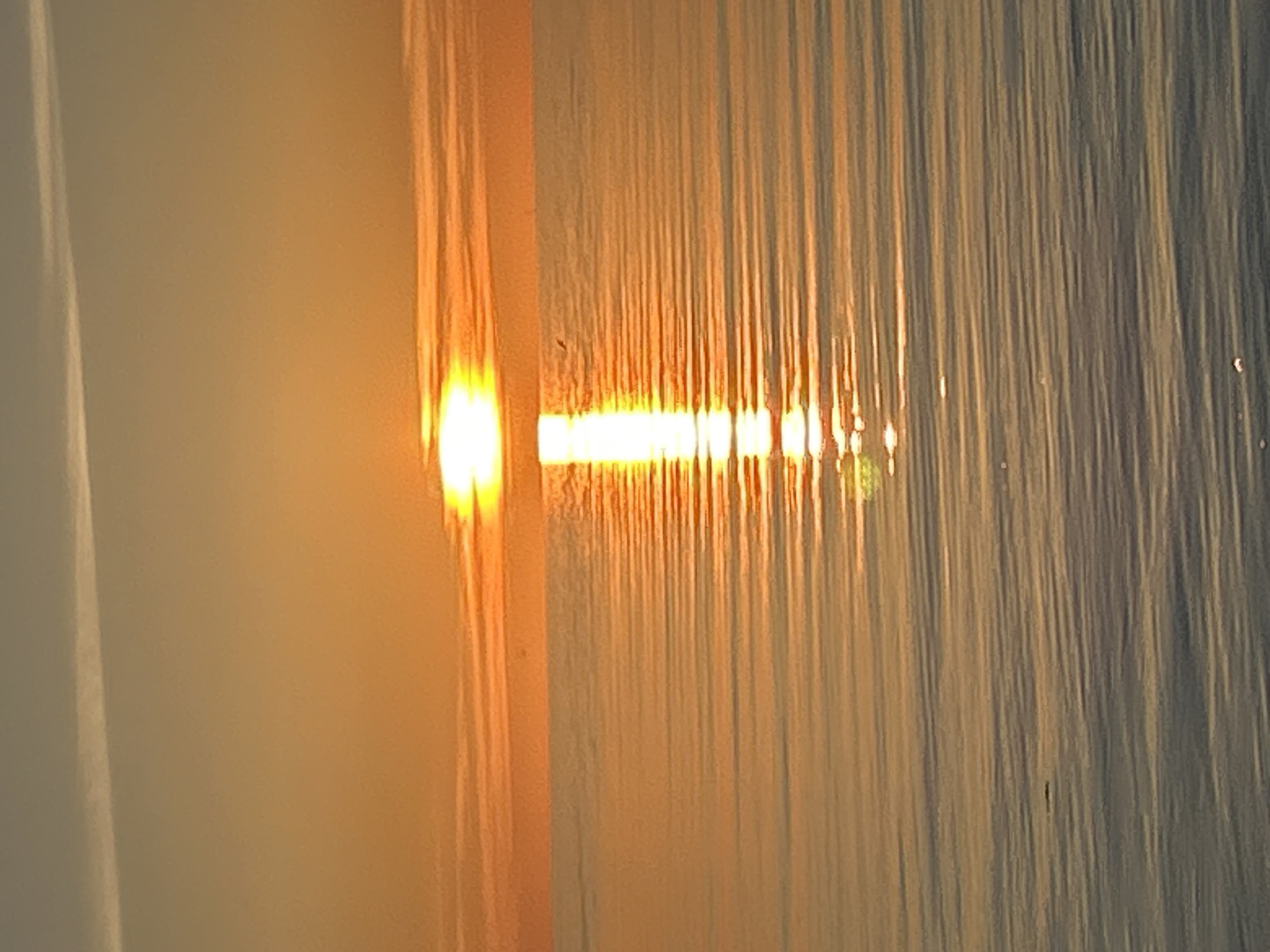 The image depicts a sunset over a body of water in Tallinn, Estonia. The upper third of the frame shows the sky, which transitions from a light blue-grey at the top to an intense orange-yellow at the horizon. A faint, thin, white linear streak, consistent with a contrail or cirrus cloud, is visible diagonally across the upper left sky. Horizontal cloud formations are present above the sun, illuminated in shades of orange.
The sun, a brilliant yellow-white light source, is positioned centrally along the horizon, casting a distinct, bright, vertical reflection path directly onto the water below.
The lower two-thirds of the image is occupied by a calm body of water, characterized by numerous parallel, horizontal ripples stretching across its surface. The water's color ranges from a vibrant orange-yellow where it reflects the sun's direct light, to a deeper, cooler blue-grey towards the foreground and areas outside the sun's reflection path. A small, dark, oblong object, approximately 1-2 pixels wide and several pixels long, floats in the lower-middle section of the water. Additionally, a minute, dark, irregular speck, possibly an airborne bird, is discernible in the sky against the orange light, slightly above and to the left of the sun's reflection on the water.