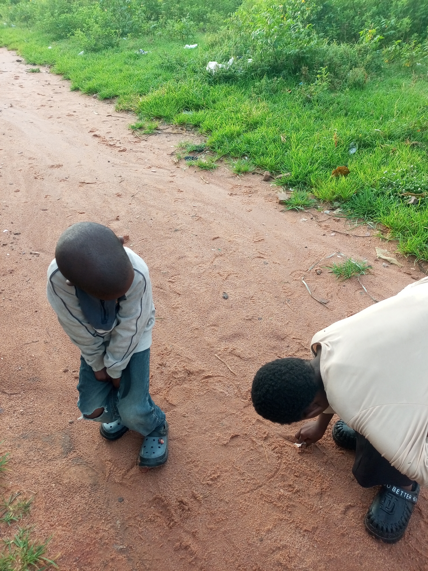 The image depicts two young boys on a dirt road in Tempure, Nigeria. The boy on the left, wearing a light grey sweatshirt and blue jeans with a tear on the knee, stands with his head bowed and hands clasped in front of him. He is wearing blue clogs. The boy on the right is crouched down, intently drawing or writing in the dirt with a stick. He is wearing a light-colored long-sleeved shirt and black pants, with black clogs on his feet that have white lettering on them. The road is a reddish-brown color and appears to be unpaved. To the right of the road, there is an area of lush green grass and dense vegetation, with some litter visible among the plants. The overall scene suggests an outdoor, possibly rural or semi-urban, environment.