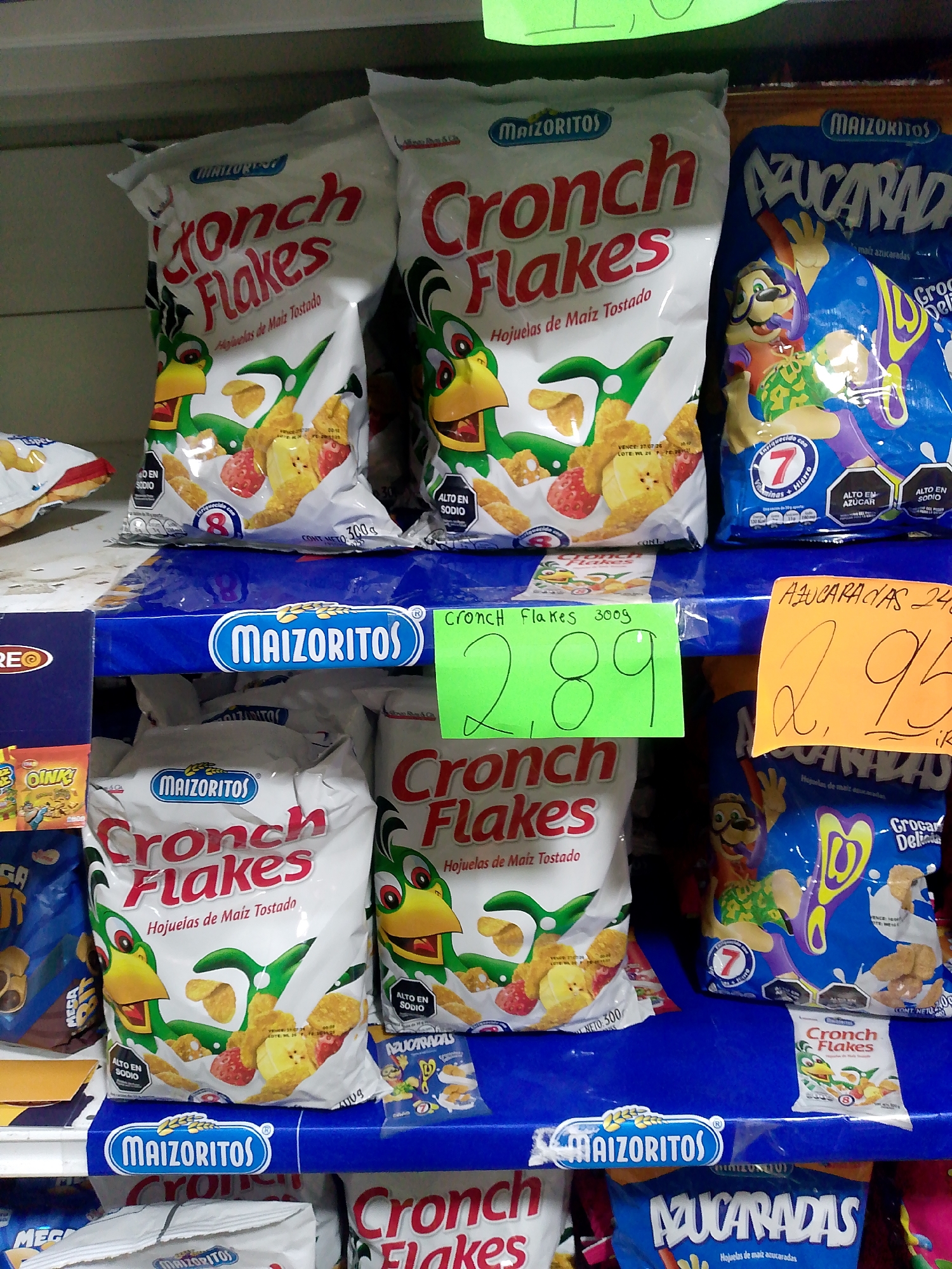 The image displays a retail store aisle in Juan Griego, Venezuela, focusing on a shelf arrangement of breakfast cereals. White "Maizoritos Cronch Flakes Hojuelas de Maíz Tostado" bags, each depicting a green cartoon bird, corn flakes, strawberries, and bananas, are prominently featured. Adjacent blue bags of "Maizoritos Azucaradas Hojuelas de maíz azucaradas" show a cartoon character with a mustache and corn flakes in milk. A green handwritten sign labels "Cronch Flakes 300g" at "2.89," while an orange handwritten sign marks "Azucaradas 240g" at "2.95." Multiple cereal bags include octagonal warning labels for "ALTO EN SODIO" (High in Sodium) and "ALTO EN AZUCAR" (High in Sugar), alongside information about fortification with vitamins and iron. The products are arranged on blue plastic shelves, with a white shelf above and other partially visible "Maizoritos" and "OINK!" branded items on the left and lower shelves. No individuals are present within the frame.