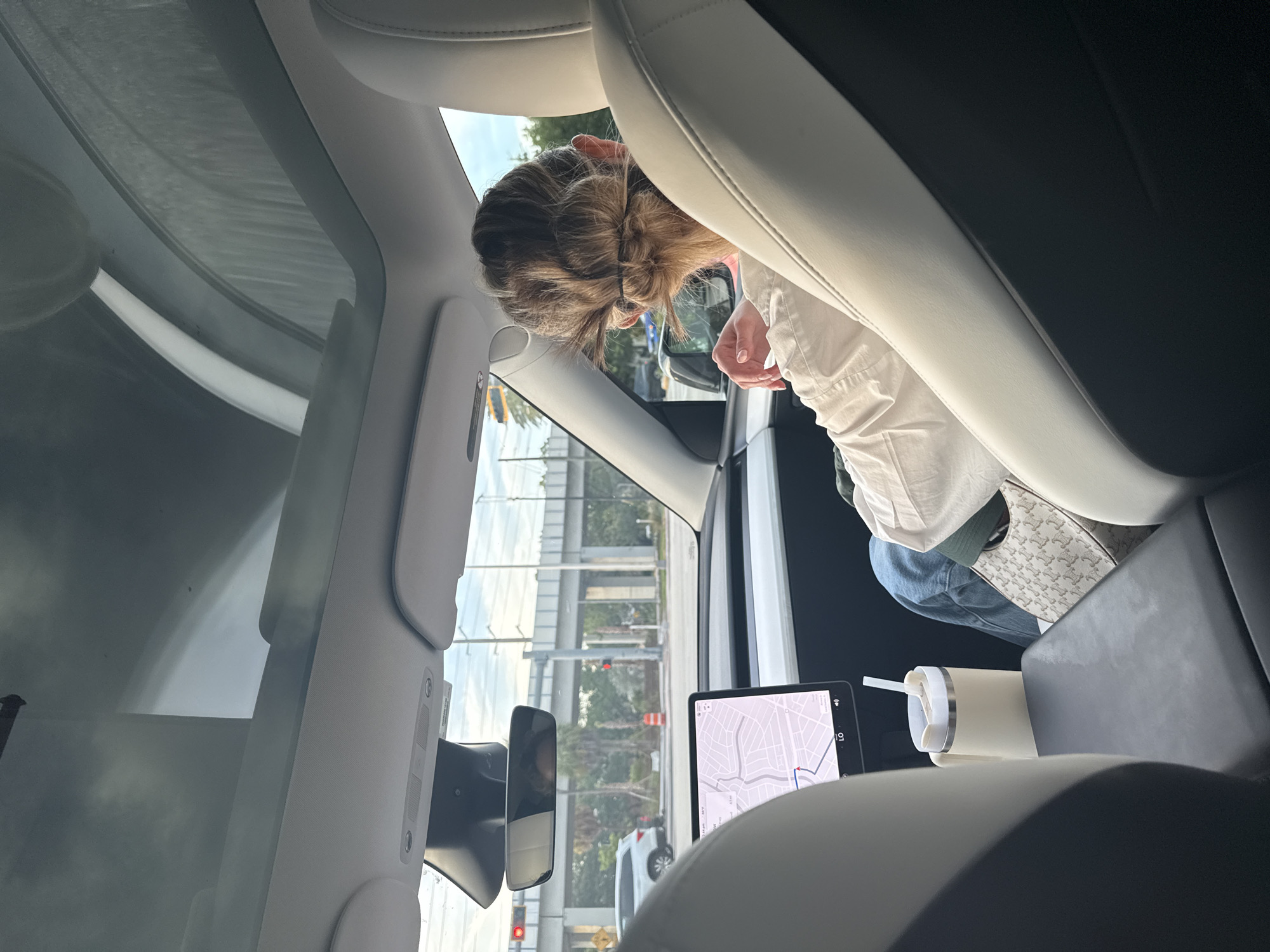 The image shows the interior of a car, likely a Tesla, with a woman's back to the camera. She has her hair pulled up in a bun and is wearing a white shirt and jeans. She is holding a beige travel mug with a straw. A car's navigation system screen is visible in the foreground, displaying a map of an urban area. Outside the car, palm trees and an overpass are visible, suggesting an outdoor setting in a city. The scene appears to be captured during daylight. The presence of a Tesla and the navigation system suggest it is a modern vehicle. The overpass and the type of vegetation hint at a location like Florida.