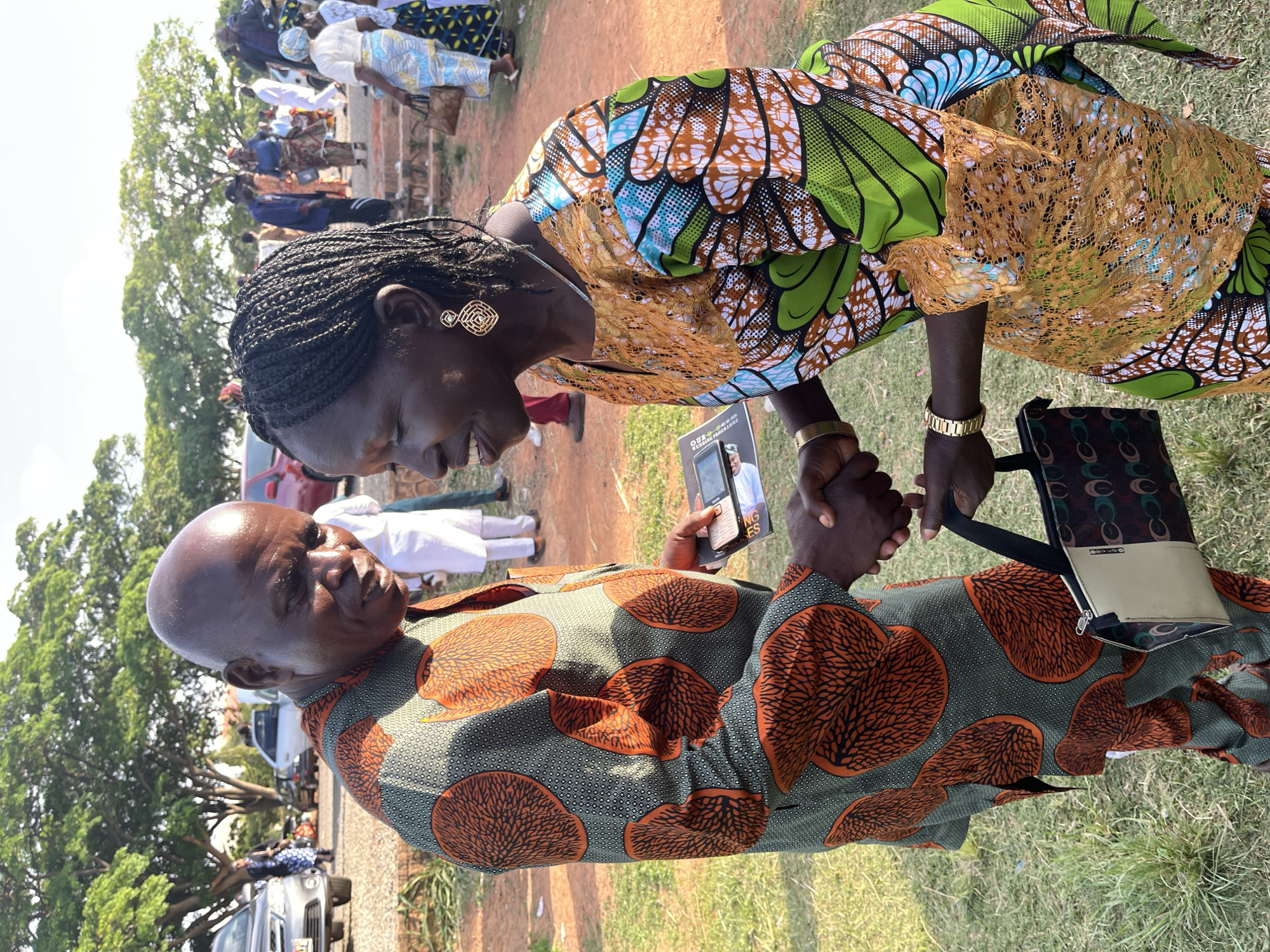 The photograph captures a scene of two individuals in Jos, Nigeria, appearing to be engaged in a solemn interaction. A bald man, wearing a green and orange patterned shirt, holds a small pamphlet in one hand and clasps hands with a woman beside him. The woman is adorned in a green, blue, and brown patterned dress with a gold lace overlay. She has her hair braided and wears gold earrings. They are standing on grass, with other people visible in the background. A handbag is slung over the man's arm. Cars and trees are visible in the background, along with other attendees dressed in various outfits, suggesting a gathering or event. The ground appears to be a mix of grass and reddish soil.