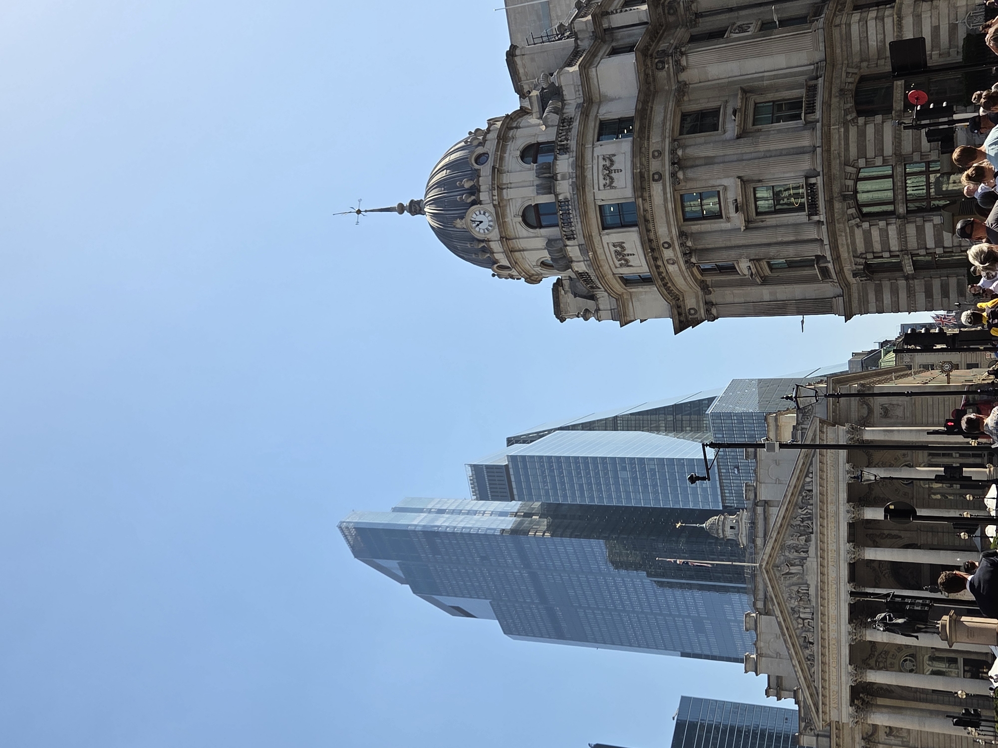 The image shows a street view in the City of London, United Kingdom. The architecture presents a mix of historical and modern elements. A classic building with a domed top featuring a clock is visible on the right. Below, a modern skyscraper with a glass facade rises. Streetlights and other street furniture are visible in the mid-ground. A crowd of people are present on the street. The sky is clear and blue.