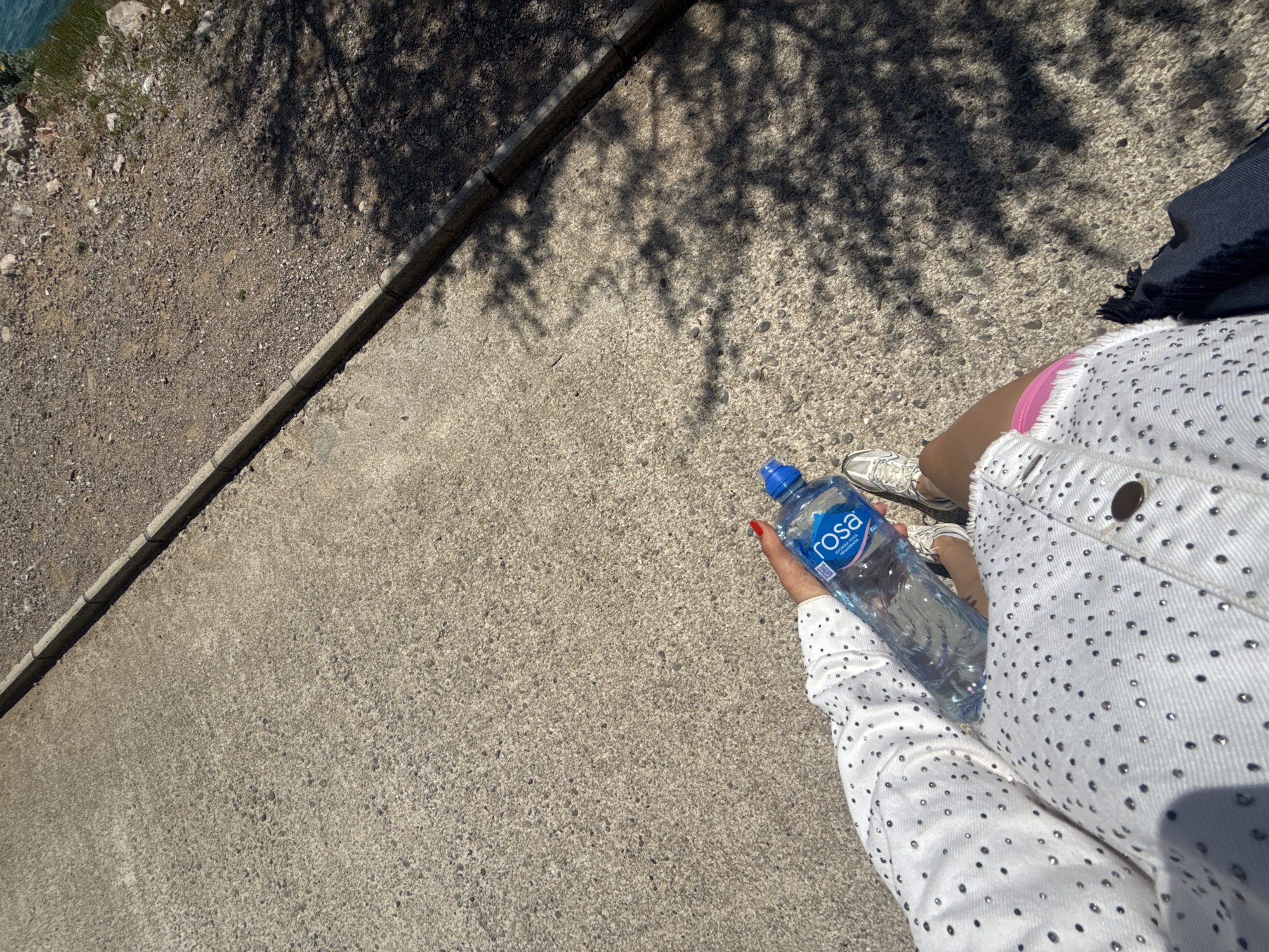 A top-down shot reveals a woman holding a bottle of "rosa" water, standing on a concrete surface bordered by gravel. Tree shadows dappled across the concrete. The woman wears a white jacket with small metallic studs, pink shorts, and white sneakers. She stands on a paved area in Unknown, Unknown, adjacent to a gravel path or shoulder.