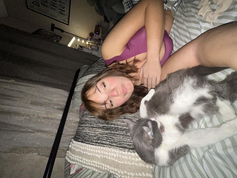 A woman in a maroon top lies on a bed beside a gray and white cat. The cat is partially covering the woman's torso. The bed has two pillows, one patterned and one striped. A metal bed frame is visible on the left. In the background, a vanity with lighted mirror and a framed poster can be seen.