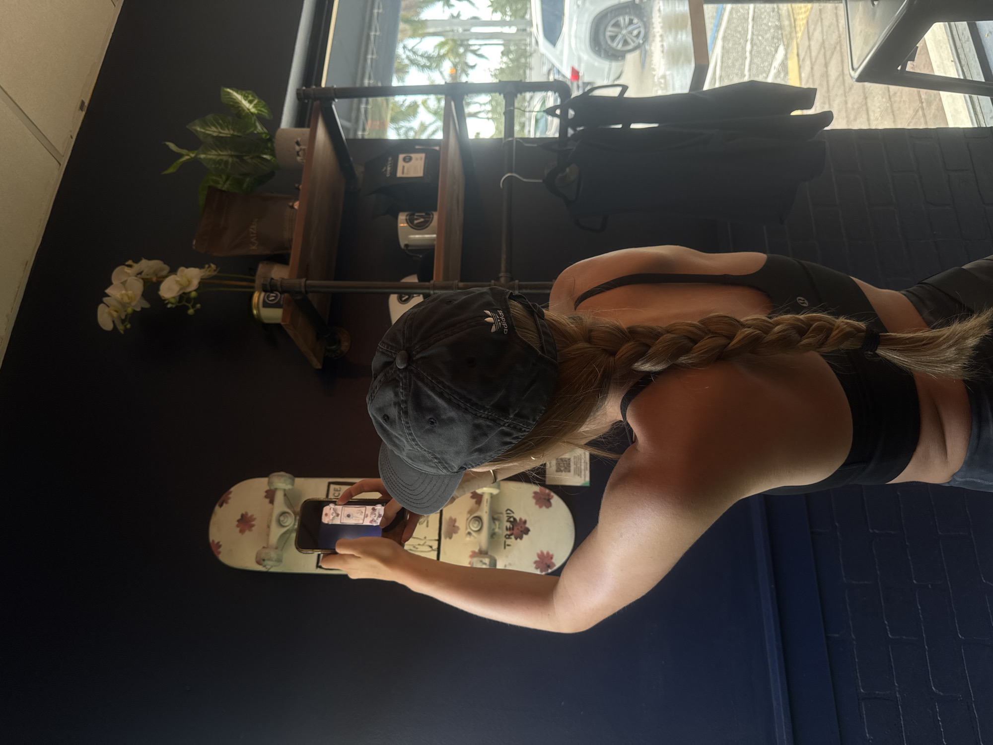 A woman in a black sports bra and an Adidas baseball cap is holding a smartphone horizontally to take a picture of a skateboard that is mounted to a dark-colored wall. The skateboard is white with a floral design and the word "TREND" written on it. In the background, there is a shelf with various items, including plants and containers. Through a window, a car is visible on a street lined with palm trees. The scene appears to be indoors, possibly in a shop or cafe. The location is likely in Boca Raton, United States of America, based on the palm trees and building style visible through the window.