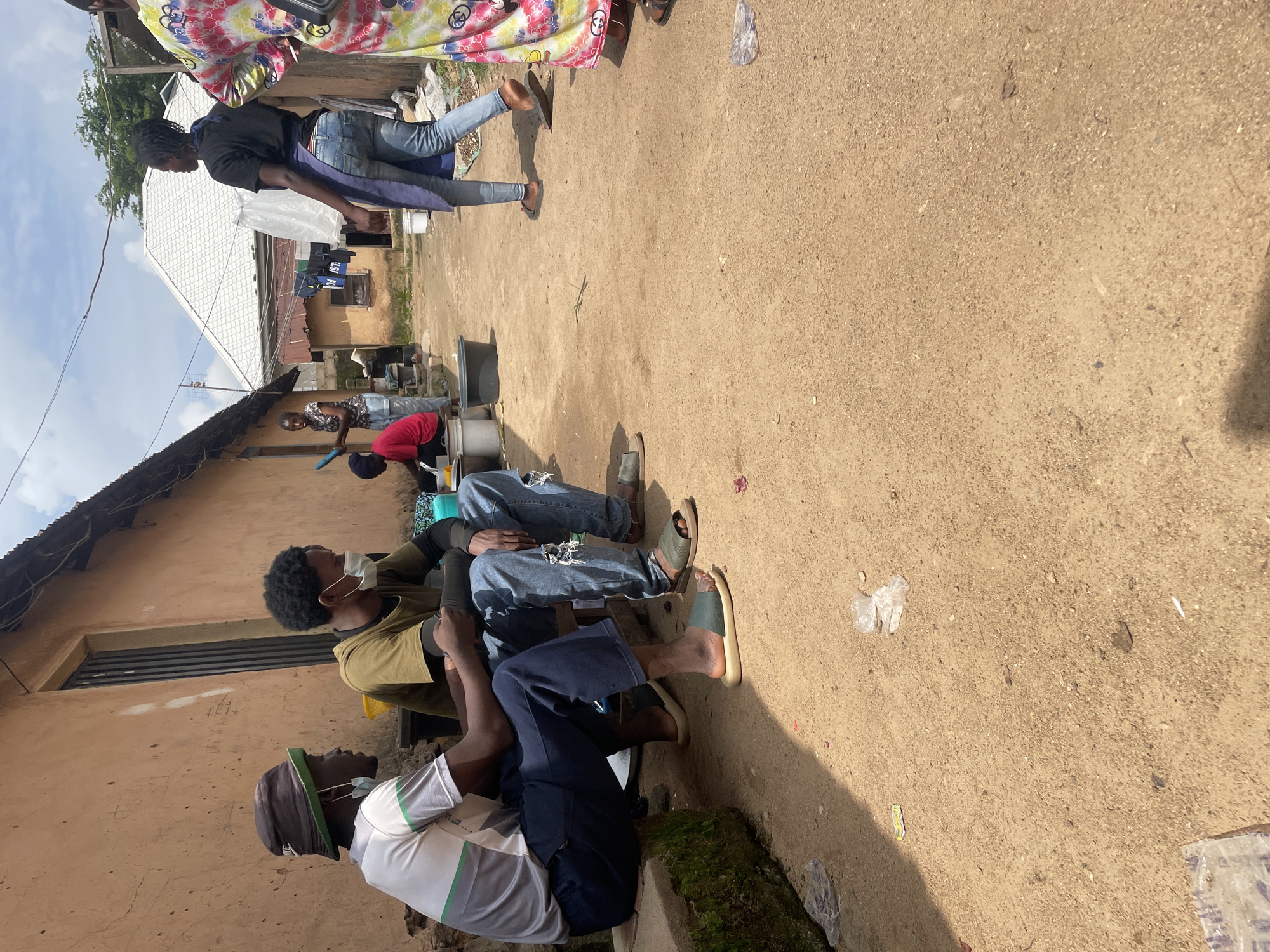 The image depicts an outdoor scene in Jos, Nigeria, likely a residential area with simple structures. In the foreground, two young men are seated on a low concrete ledge. The man on the left wears a white t-shirt with green and grey stripes, dark trousers, and a bucket hat. He has a disposable face mask hanging below his chin. The man on the right wears a dark t-shirt, ripped blue jeans, and is also wearing a disposable face mask. Both men are looking towards the left of the frame and appear to be in conversation.
Behind them, a dirt pathway leads into the background. Several other individuals are visible, engaged in various activities. A woman in the middle ground, wearing a black t-shirt and blue jeans, walks away from the camera, her back to the viewer, and has a light-colored apron over her jeans. To her left, a woman in a red t-shirt is crouching near a large grey plastic basin, possibly doing laundry or washing. Further in the background, laundry is hanging on a line. The buildings appear to be constructed from earth or mud-brick, with corrugated metal roofing. The ground is covered in loose soil and gravel, with some scattered debris. The sky is partly cloudy, suggesting it is daytime. The overall impression is of a community setting where daily life activities are taking place.