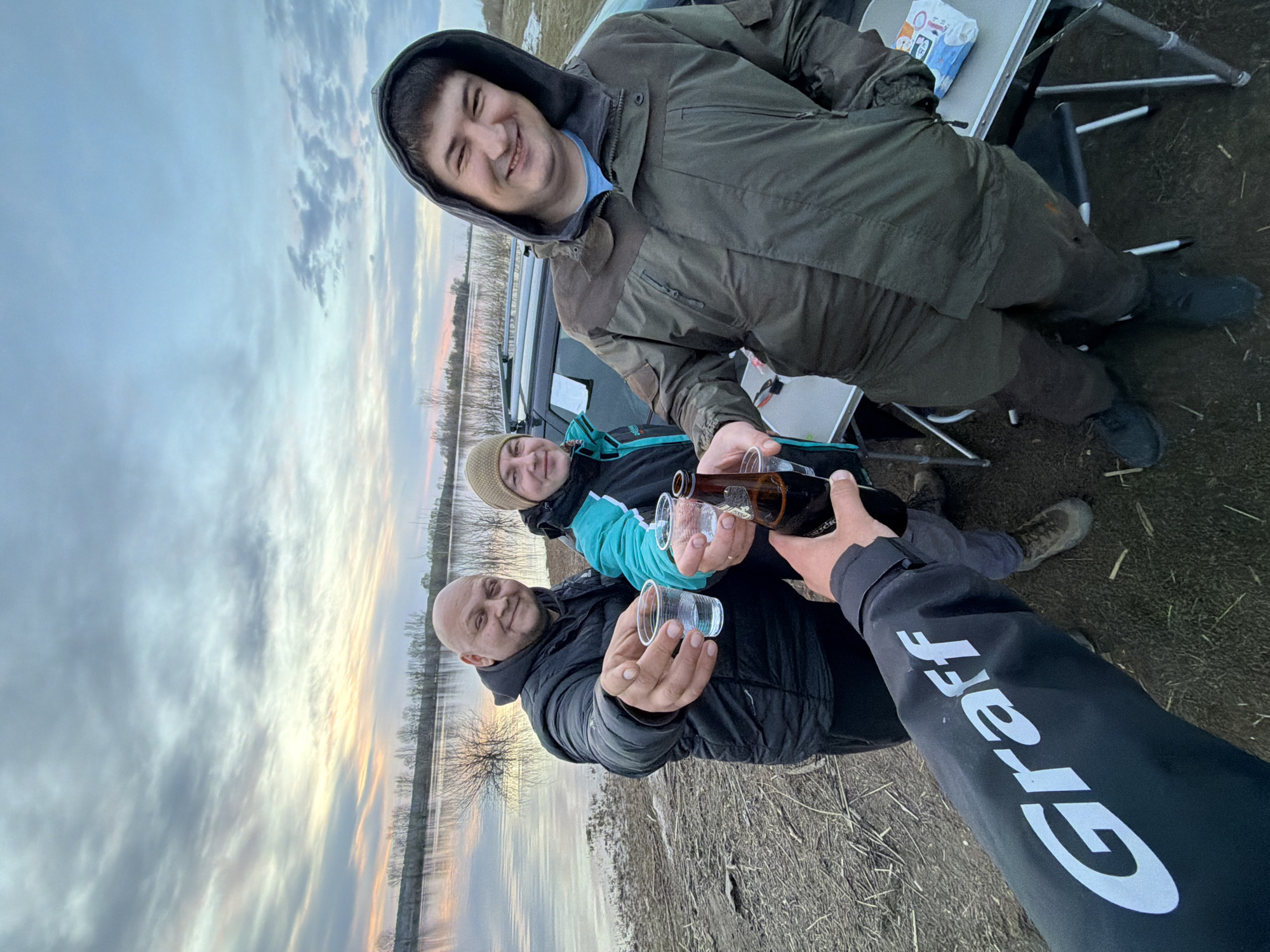 Four people are gathered in an outdoor setting. The scene appears to be near a body of water or a flooded area, as evidenced by the water visible in the background under a cloudy sky. Three individuals are visible from the waist up, while only an arm is seen of the fourth. All are holding plastic cups, and one person is pouring a brown beverage, likely beer, from a brown bottle into the cups.
The person doing the pouring is wearing a black jacket with the word "Graff" printed in large white letters on the sleeve. One of the people holding a cup is a bald man wearing a dark-colored puffer jacket. Beside him stands another person in a light-colored hat and a blue jacket. Behind them stands a man in a dark green hooded jacket. In the background, there is an SUV and a folding table with a small white box. The ground appears to be a mix of mud and dry grass.
The location is Nikolo-Berezovka, Russia.