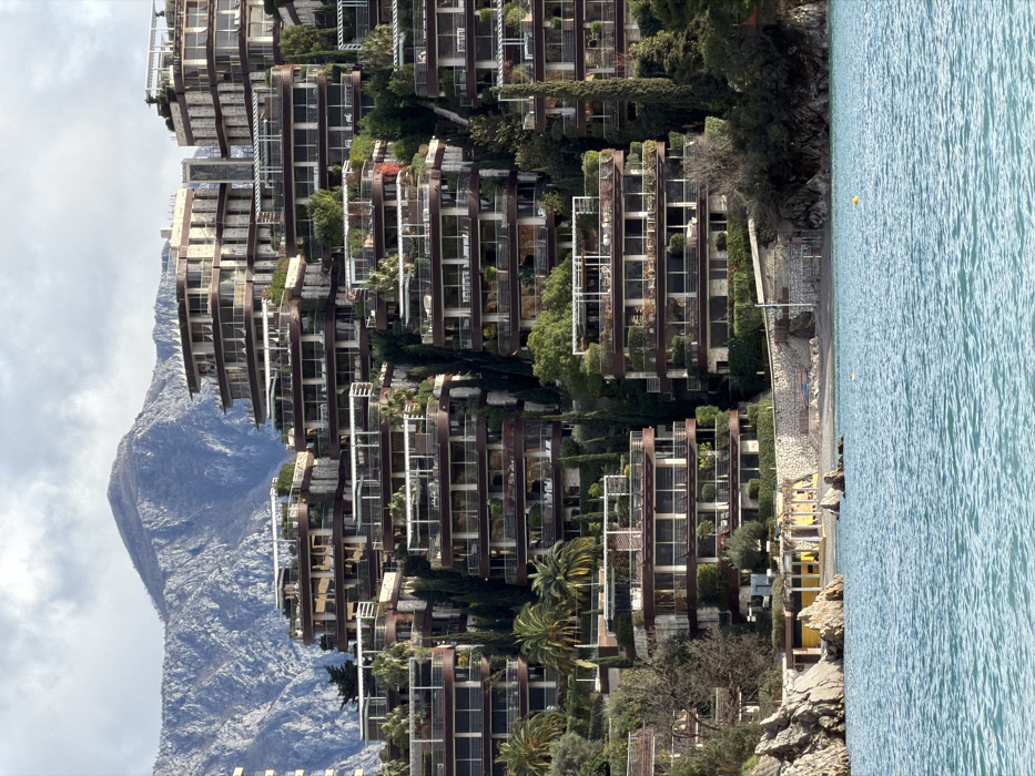This image depicts a coastal hillside scene in Boreti, Montenegro. The foreground features calm, turquoise-blue water with minimal ripples, containing two yellow spherical buoys. A narrow, light-colored gravelly beach occupies the immediate shoreline, bordered by a low stone retaining wall on the right and natural rock formations on the left. A small, single-story yellow building with striped awnings is visible on the beach, near the center-left.
Rising steeply from the beach are numerous multi-story, terraced buildings, likely residential or hotel complexes, built directly into the hillside. These structures are characterized by their light-colored facades, dark brown horizontal bands indicating balconies or rooflines, and extensive glass windows and sliding doors. Each terrace and rooftop is heavily vegetated with various plants, including small trees, shrubs, and occasional palm trees. Tall, slender, dark green cypress-like trees are interspersed vertically throughout the complex, accentuating the hillside's contours. The buildings exhibit an irregular, cascading architectural style, with curved and angular sections.
In the background, a large, rugged mountain range with visible grey rock faces and sparse vegetation rises under a partly cloudy sky. No individuals are visible in the scene.